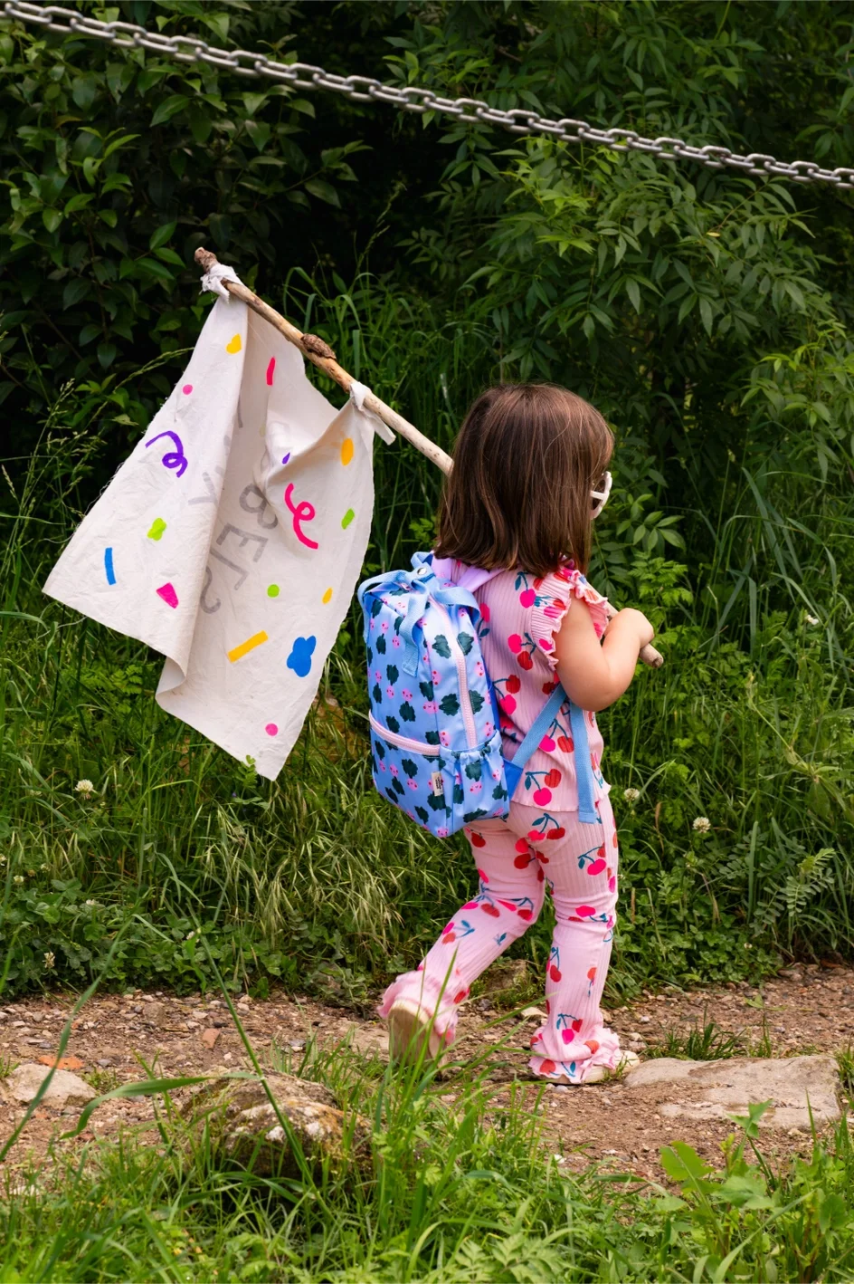 A young girl walking along a dirt trail in a lush green area, carrying a blue backpack with dark blue clouds, wearing pink pajamas with cherries, and holding a stick with a cloth attached decorated with colorful shapes.