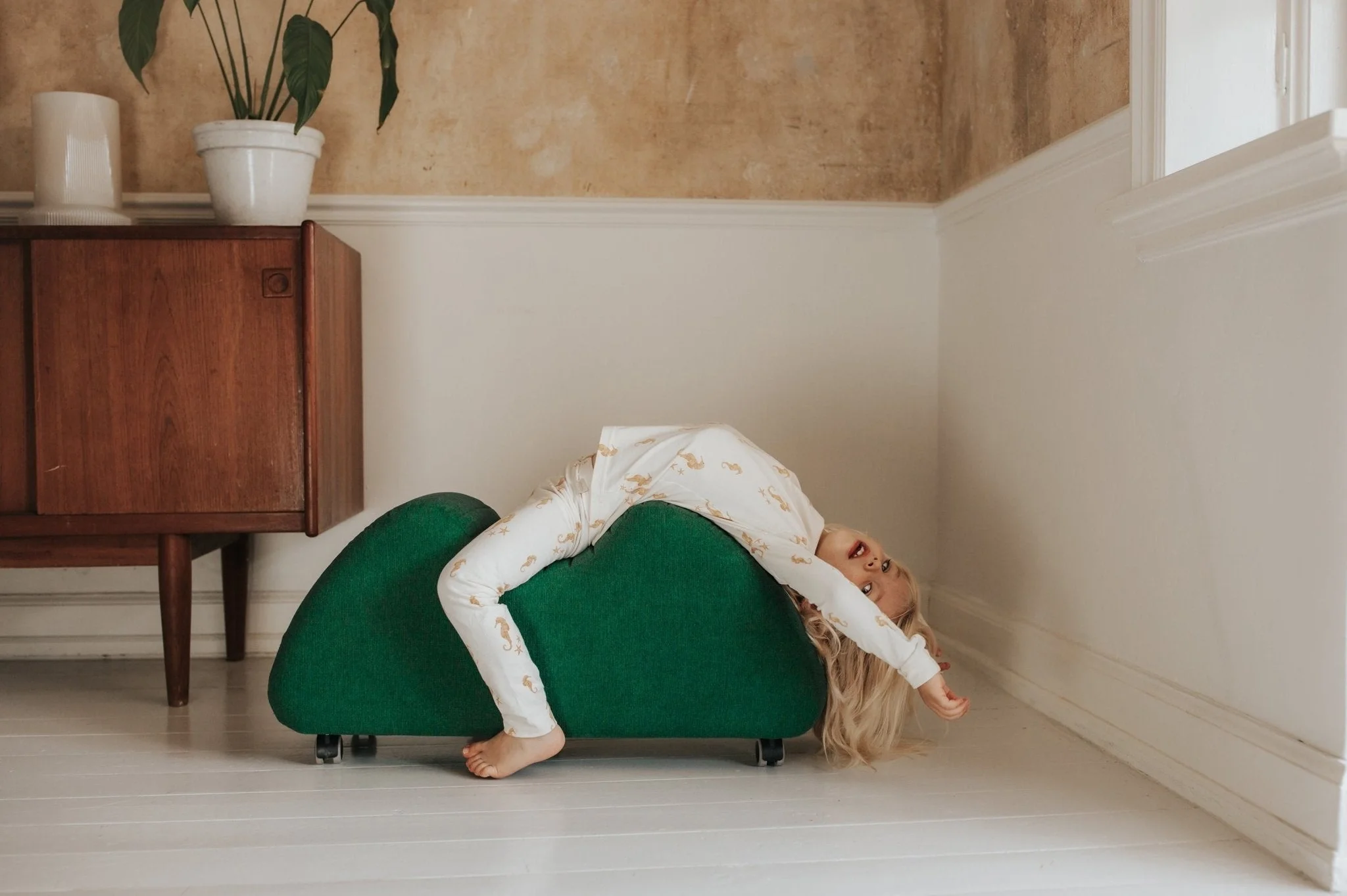 A young girl with blonde hair lying upside down on a green couch in a room with beige and white walls, next to a wooden cabinet with potted plants on top.