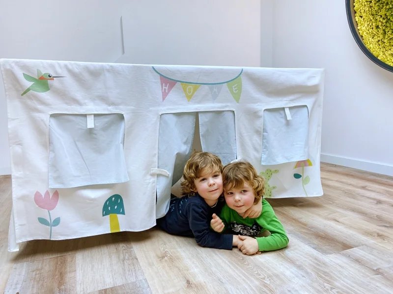 Two young boys are lying on the floor inside and in front of a white fabric play tent decorated with colorful illustrations and the word "HOME" on a banner.