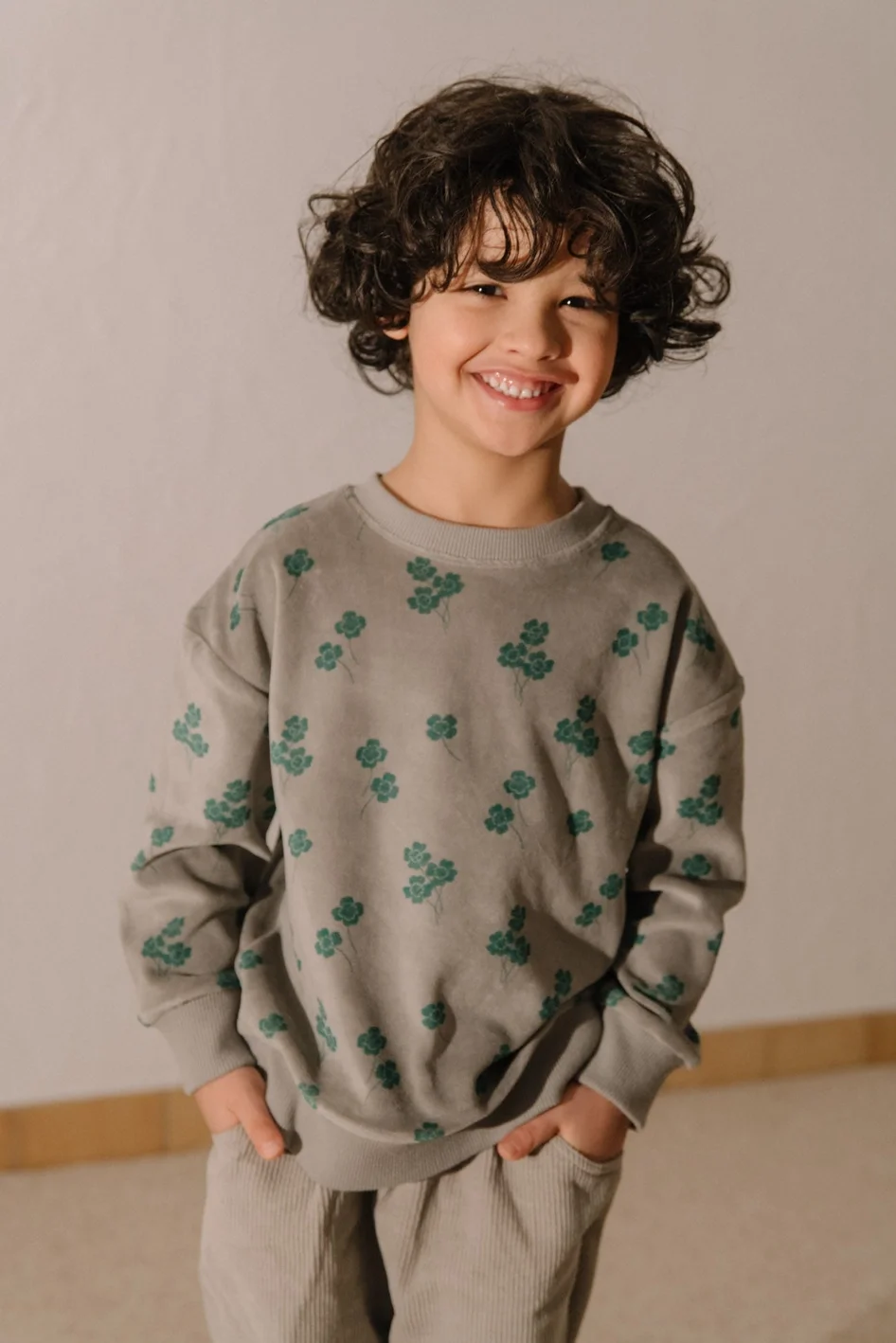 A young boy with curly brown hair and a big smile, wearing a gray sweatshirt with green floral patterns and light-colored pants, standing indoors against a plain wall.