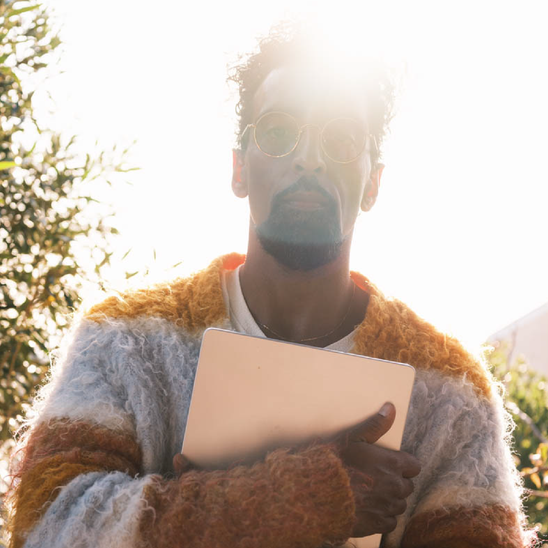 A man with sunglasses and a beard holding a book outdoors, with bright sunlight behind him and trees in the background.