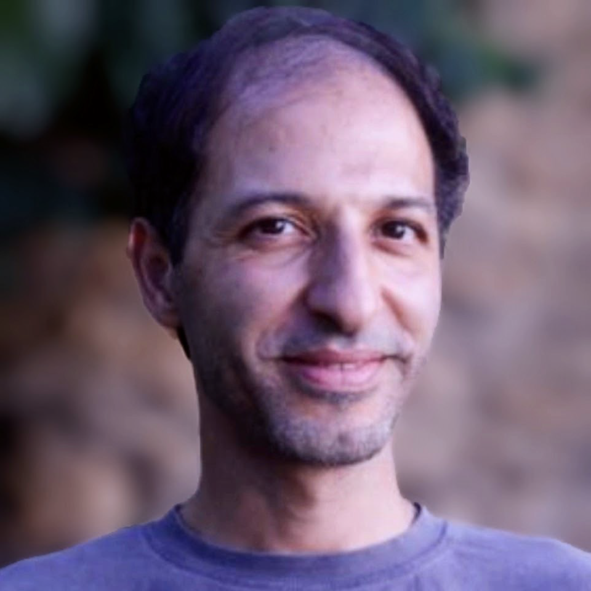 Close-up of a smiling man with short dark hair, light skin, wearing a gray t-shirt, outdoors with blurred background.