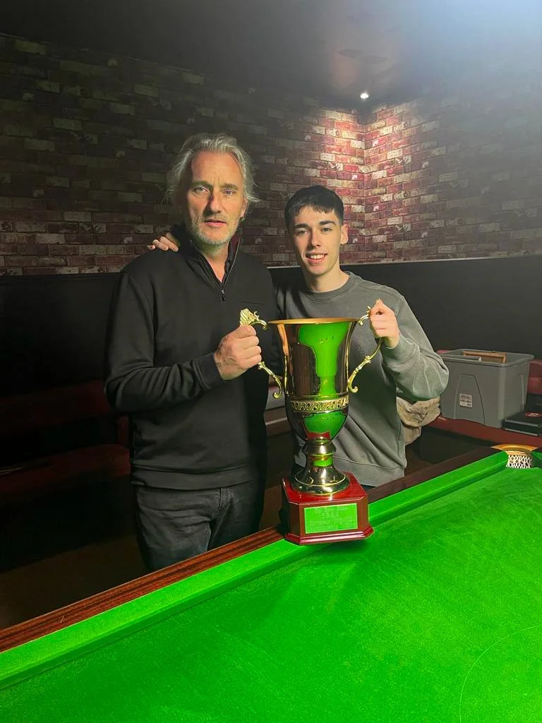 Two men standing by a pool table, holding a large gold trophy.