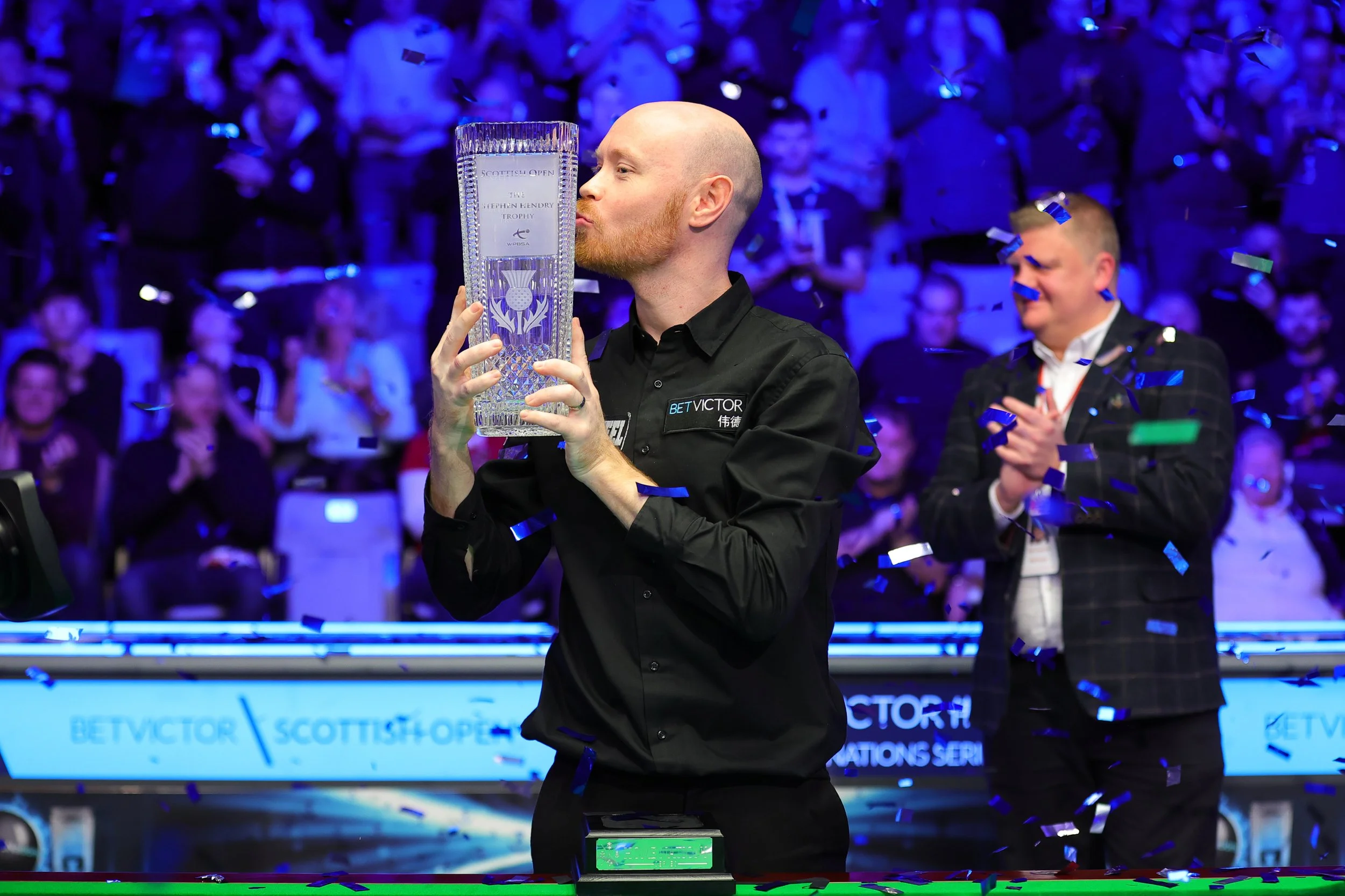 A man kissing a trophy at a pool game tournament, with an audience clapping and celebrating in the background, and blue confetti falling.
