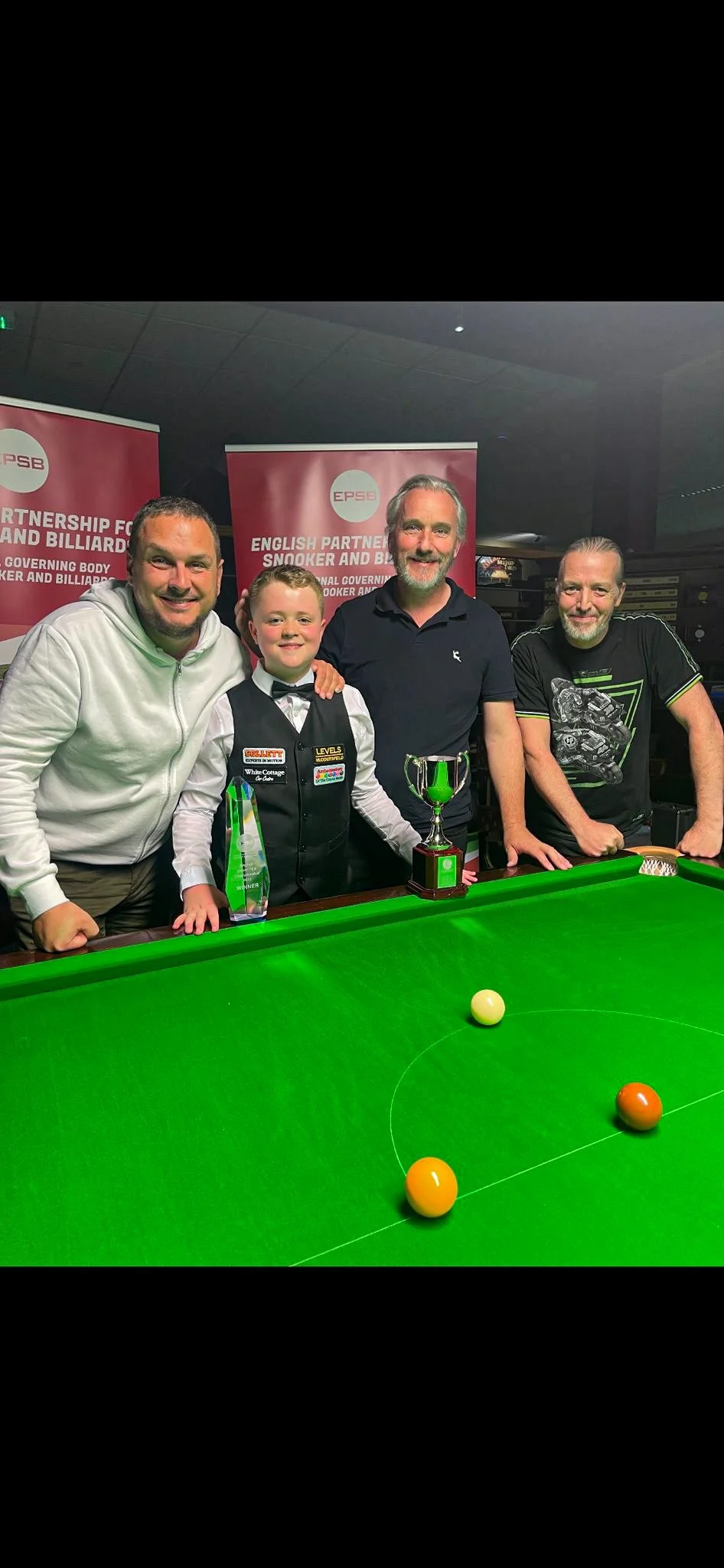 Four men and a boy standing behind a pool table with snooker balls, trophies, and awards, in front of banners for the English Partnership for Snooker and Billiards.