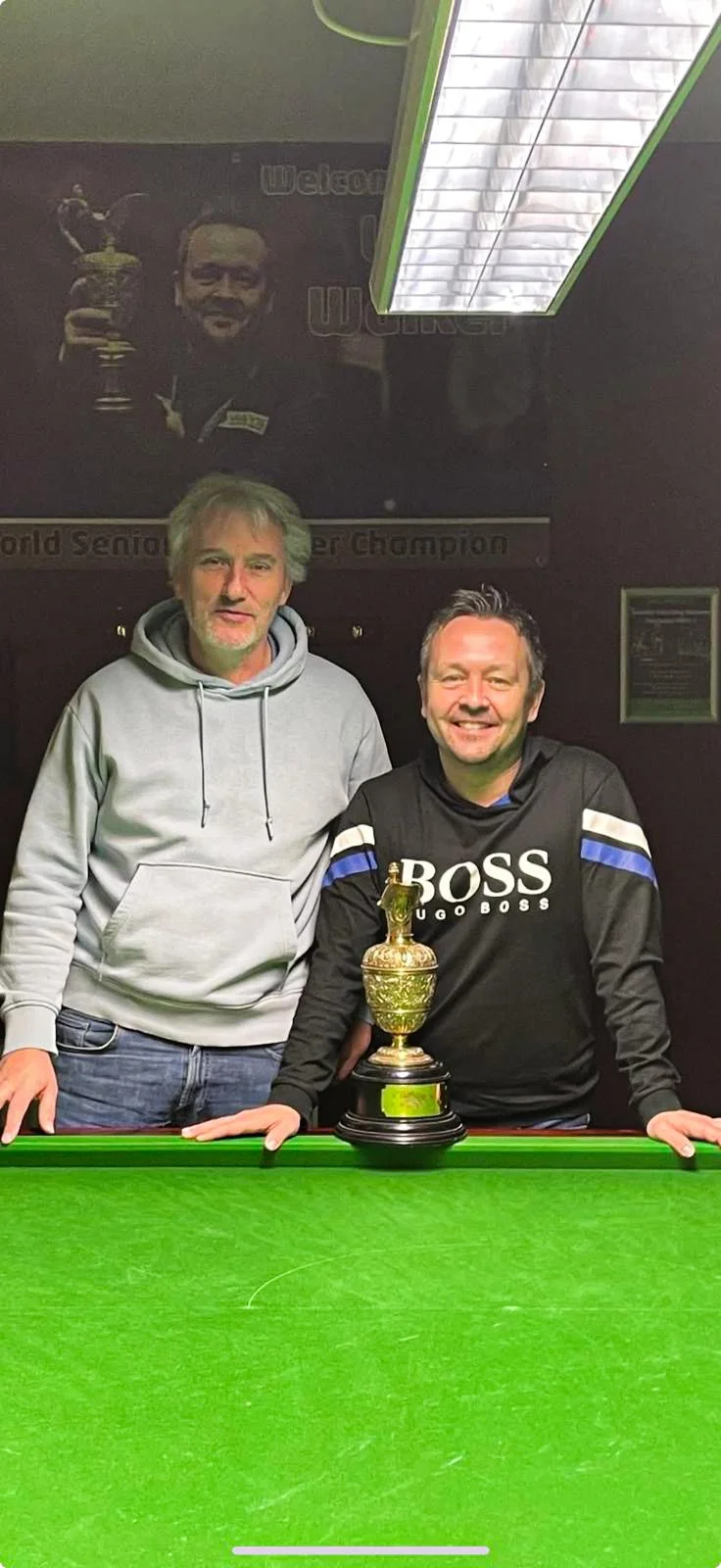 Two men standing behind a pool table with a trophy in front of them. One man is wearing a grey hoodie and the other a black Hugo Boss sweatshirt. In the background, a large poster of a man holding a trophy is visible.