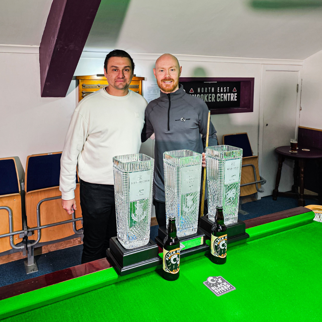 Two men standing behind a snooker table with trophies and bottles. One man is dressed in white, the other in a gray hoodie. There is a sign reading 'North East Snooker Centre' in the background.