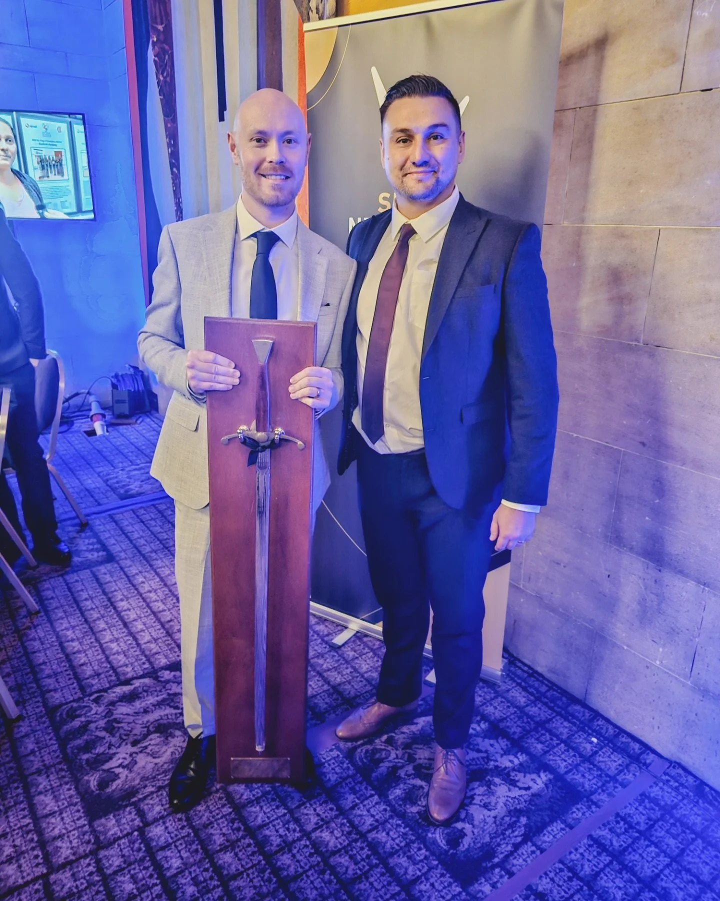 Two men in suits standing together at an event, one holding a large wooden sword award.