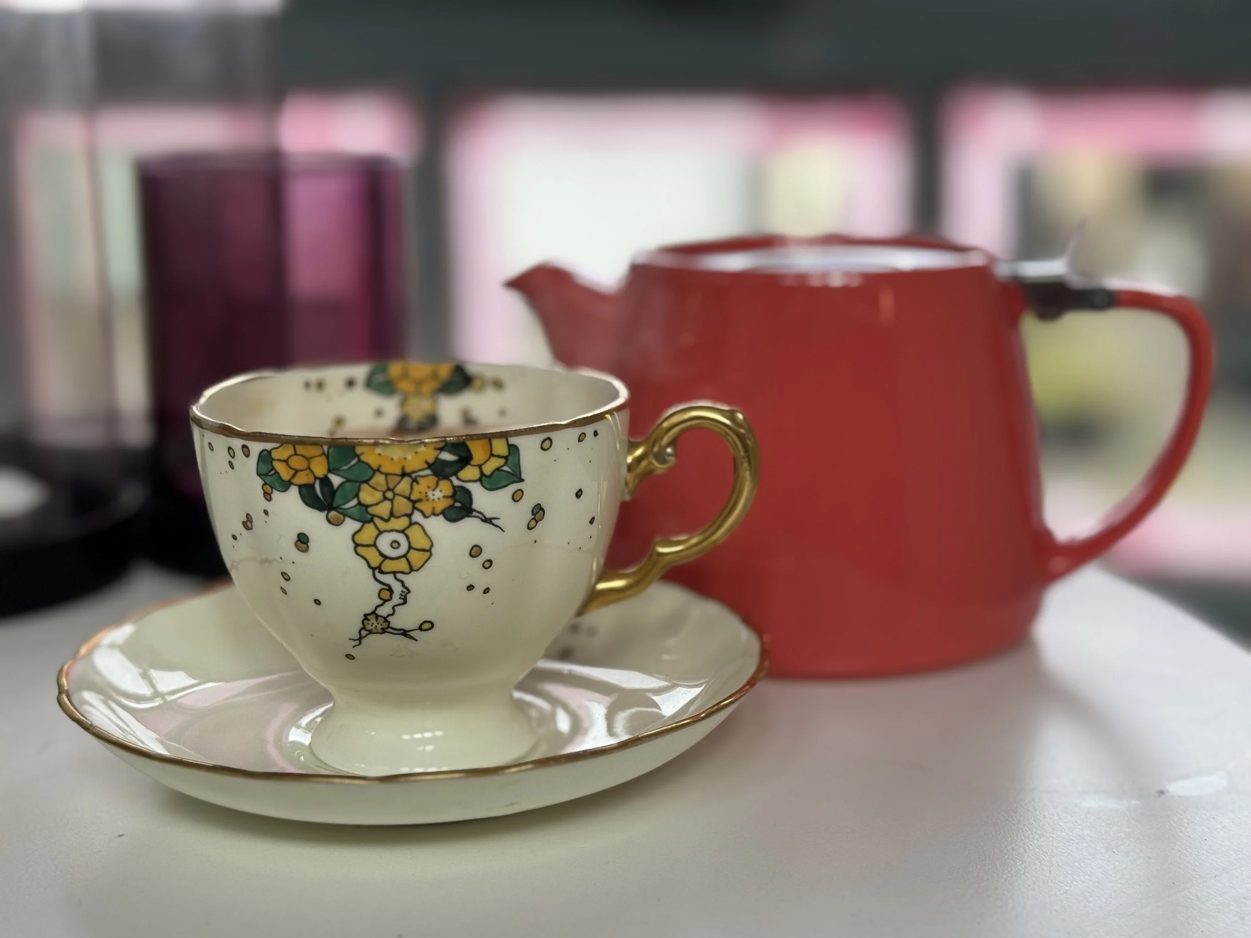 A vintage decorated china teacup and saucer sit in front of a contemporary red teapot