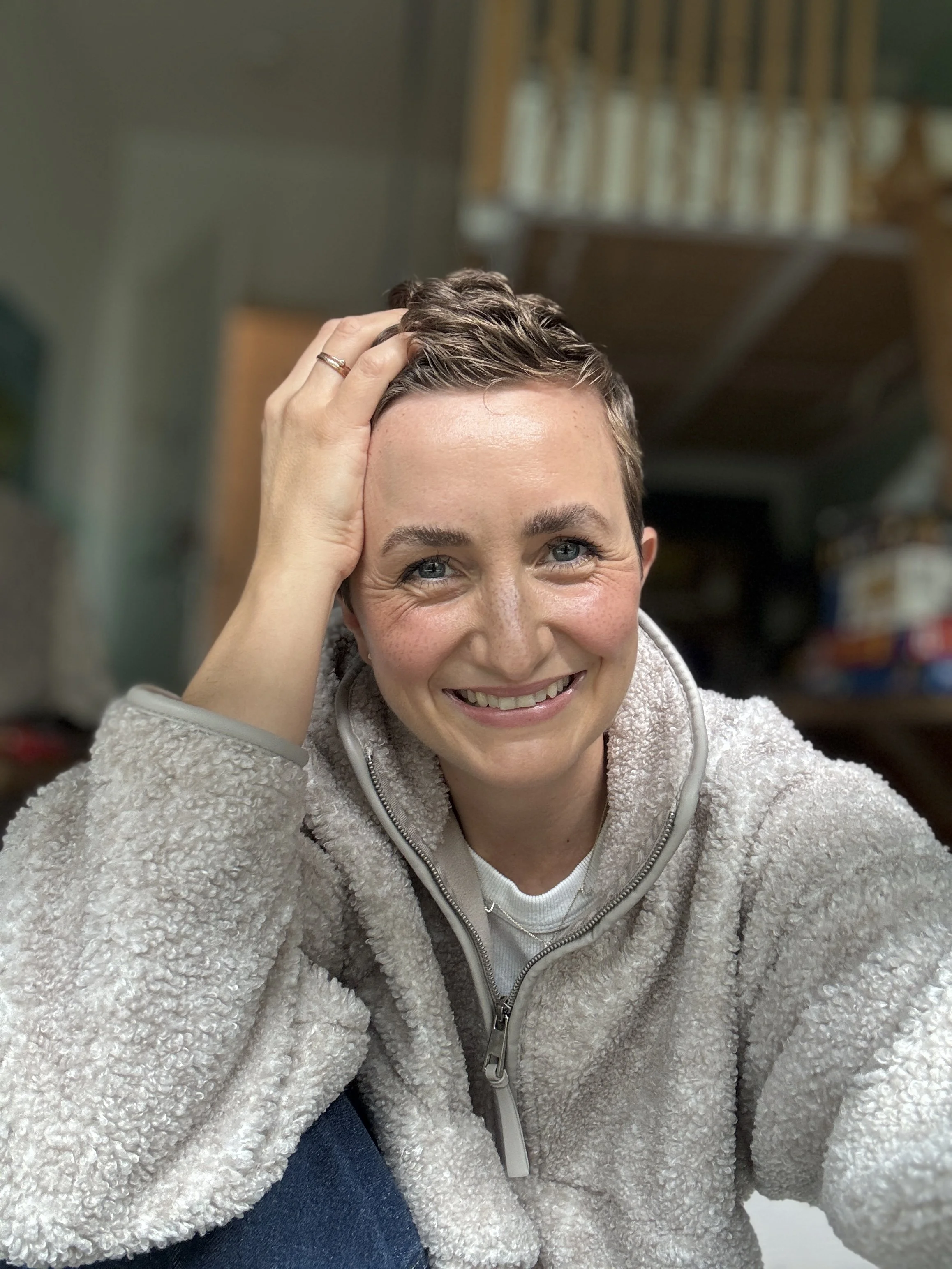 Eine lächelnde Frau mit kurzen braunen Haaren trägt ein helles, flauschiges Oberteil und sitzt in einem Raum mit einem Holzdach und Buchregalen im Hintergrund.