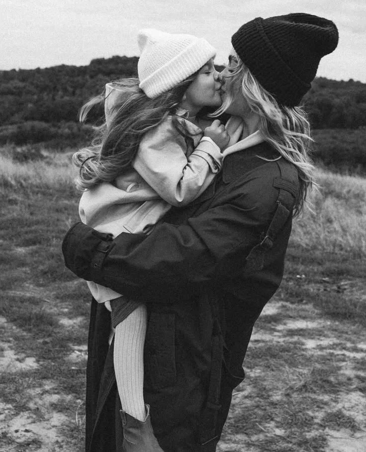 Zwei Frauen, eine ältere und eine jüngere, küssen sich im Freien bei Sonnenuntergang, beide tragen warme Mützen, die jüngere hält die ältere in den Armen.