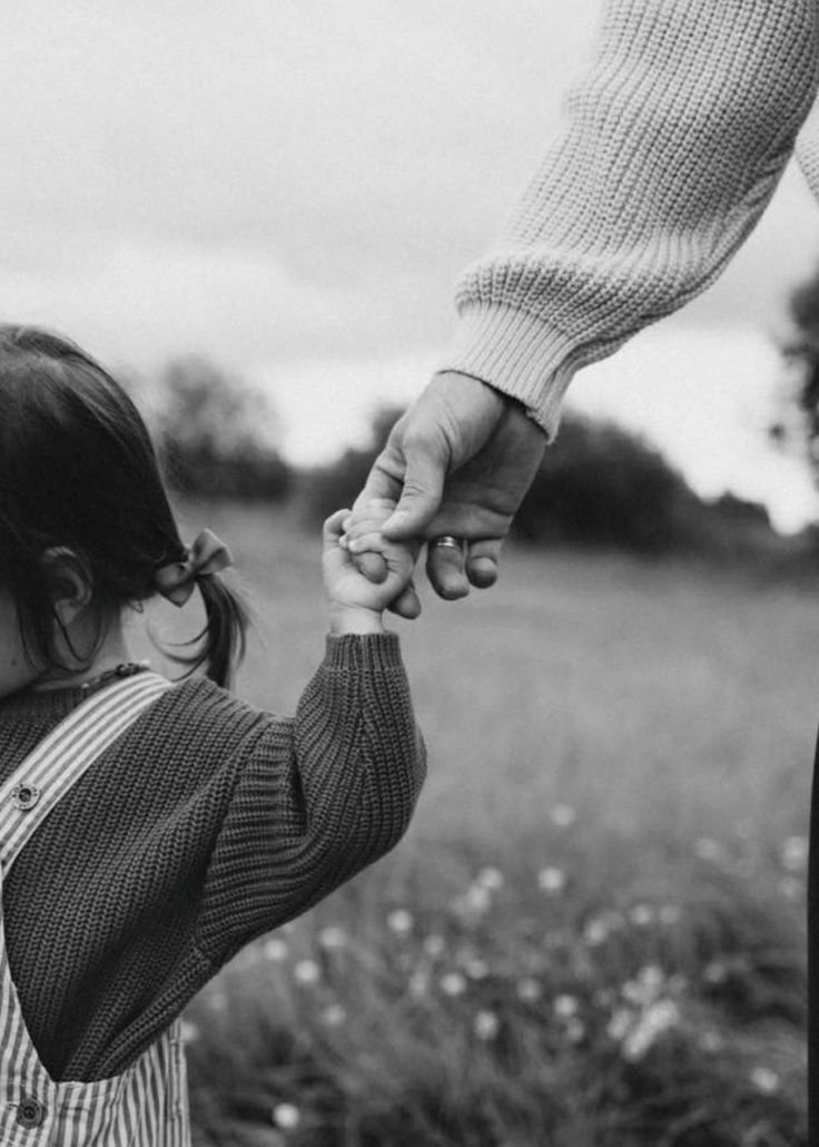 Ein Erwachsener hält die Hand eines Kindes in einem Feld bei Tageslicht, Schwarz-Weiß-Foto.