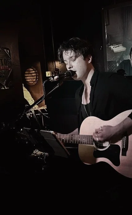 Junge Person spielt Gitarre und singt in einem dunklen Raum.