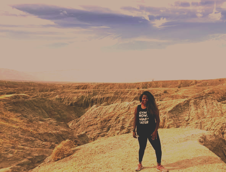 A woman standing on rocky terrain in a desert landscape with dramatic cliffs and a partly cloudy sky.
