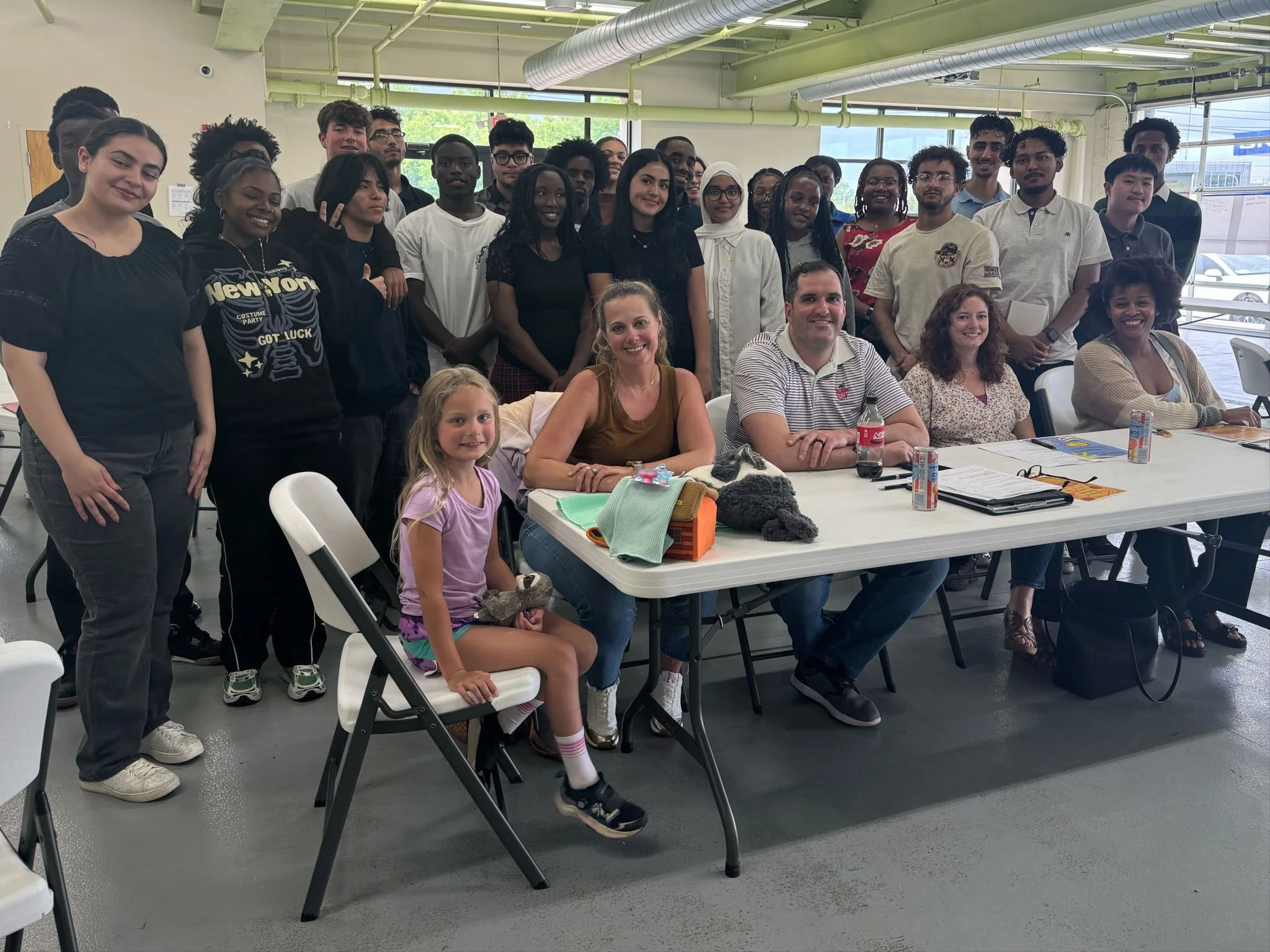 Group photo of diverse people in a classroom or meeting room, with some seated at a table and others standing behind, smiling at the camera.