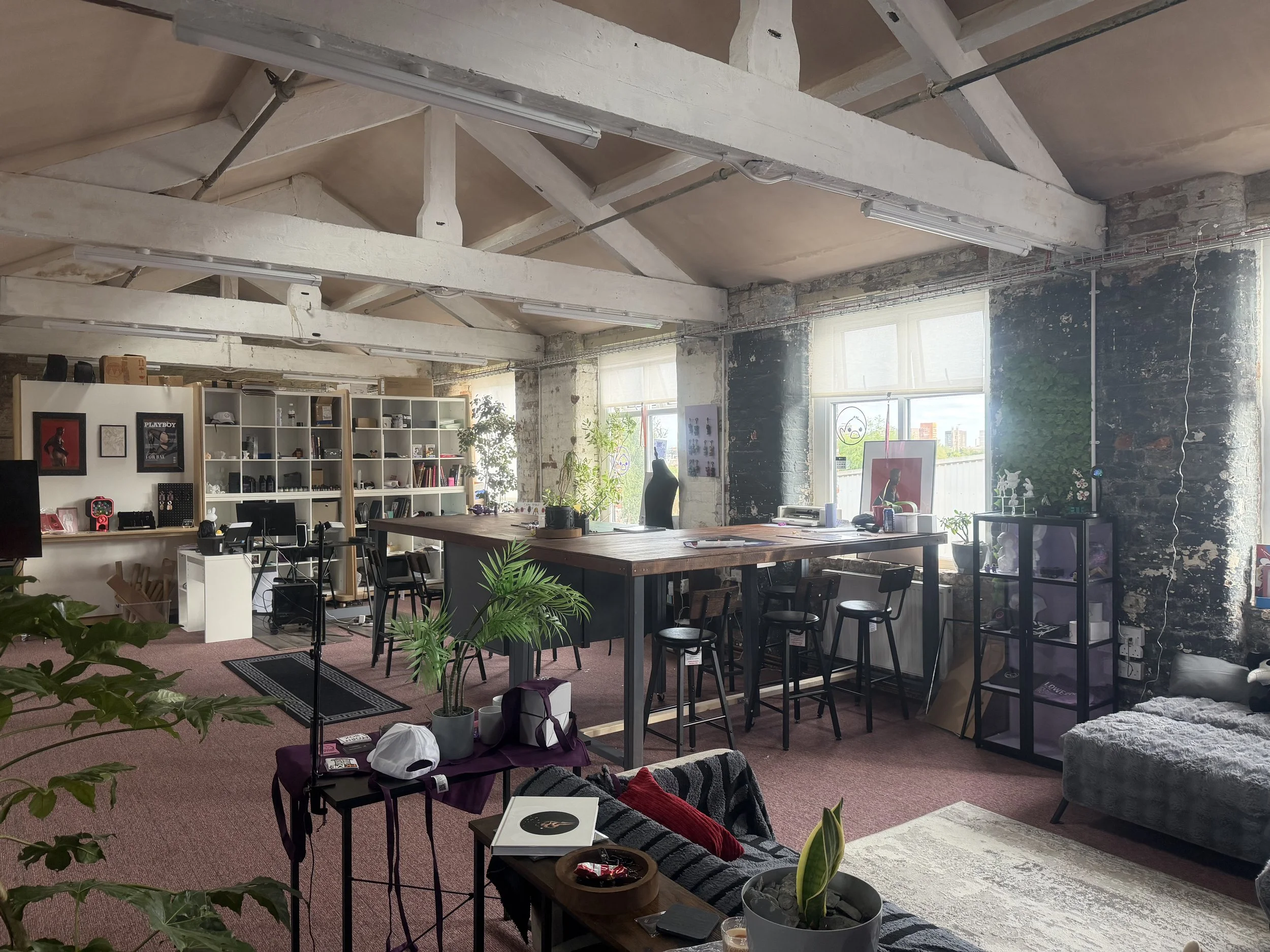 A spacious, industrial-style office with exposed concrete beams and walls, large windows letting in natural light, a variety of plants, and furniture including tables, chairs, and shelves with decorative items.