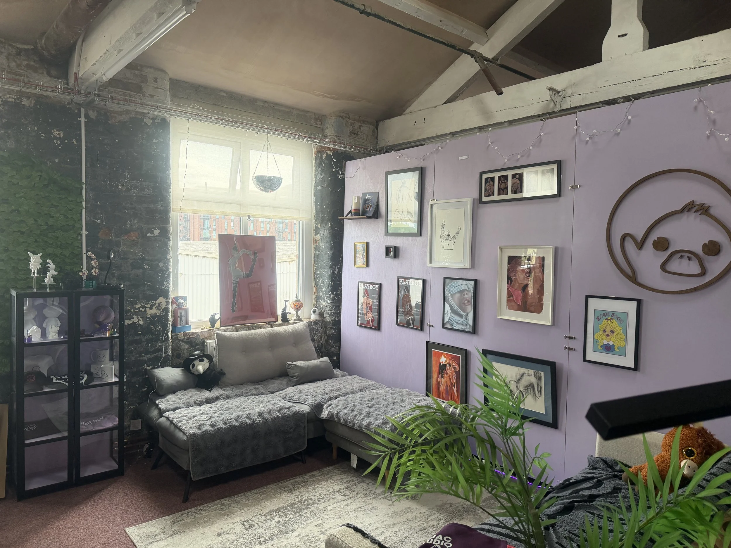 Living room with purple wall decorated with framed artwork and a large wooden logo. There is a window with blinds allowing natural light, orbs string lights, a gray couch with pillows, a black display case with figurines, and a plant.