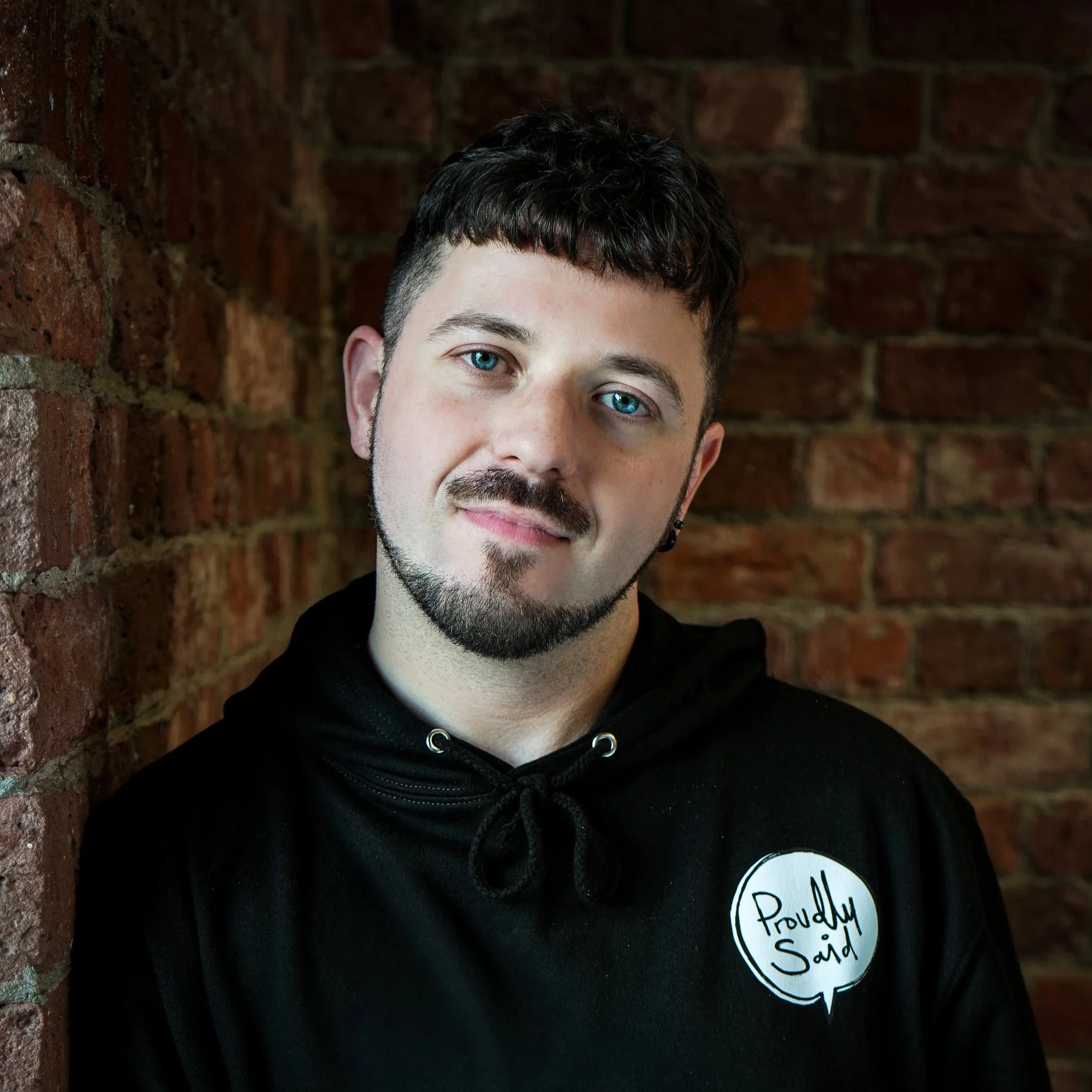 Close-up of a young man with curly dark hair, blue eyes, and facial hair, standing against a brick wall. He is wearing a black hoodie with a circular badge that reads 'Proudly Said' and has a serious expression.