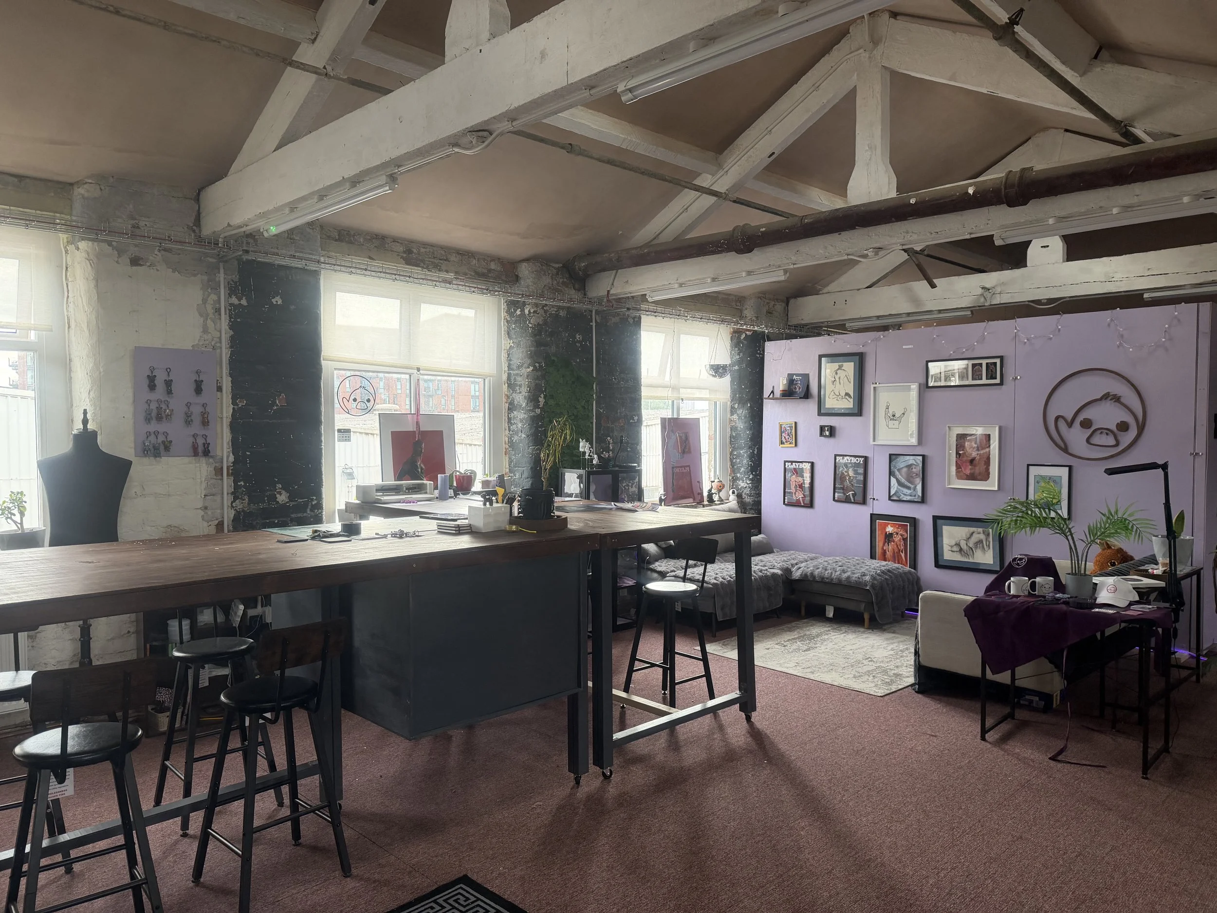 An indoor art gallery or studio space with exposed beams, large windows, and a purple wall decorated with framed artwork, a wall sculpture, and decorative items. There are tables, chairs, and plants in the room.