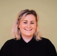 Kvinne med blond, skulderlangt hår, smilende, iført svart topp, mot en lysfarget bakgrunn. Kristine Tingvik Aas