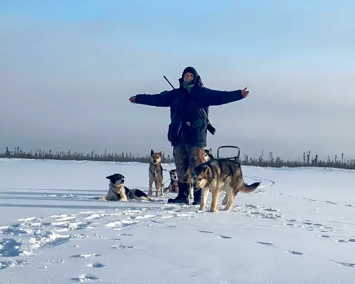 Person with four dogs standing on snow-covered landscape with arms outstretched and a backdrop of sky and distant trees.
