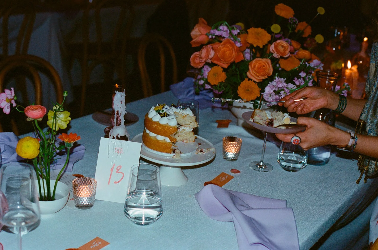 A celebratory table setting with a partially sliced cake, floral arrangements, candles, glasses of water, and a birthday candle on the cake.