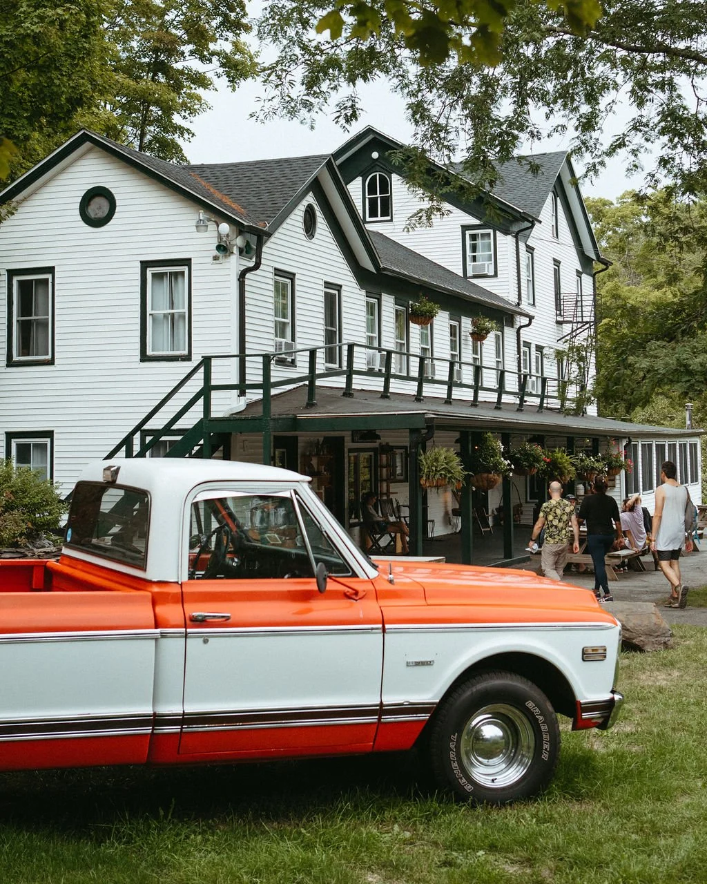 A white, three-story house with black window frames and a porch with hanging plants. An orange and white vintage pickup truck is parked in the front yard. People are walking around and sitting on the porch.