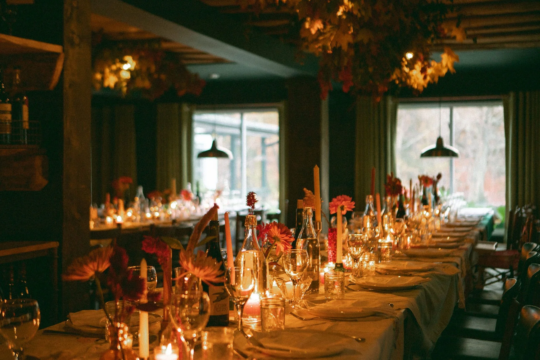 A warmly lit dining room with a long table set for a celebration, adorned with candles, flowers, and glassware, with windows showing an outdoor autumn scene.