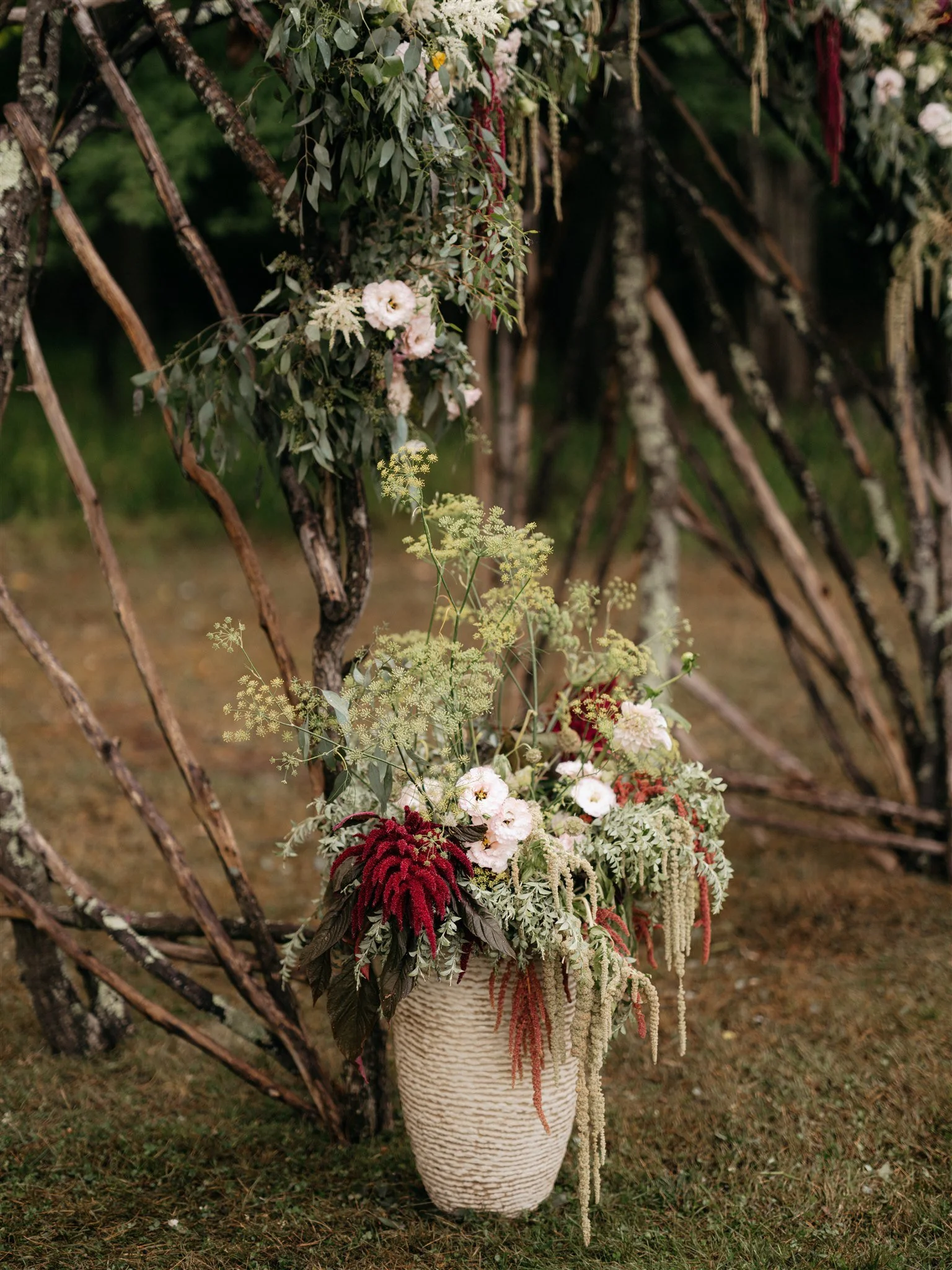 A large floral arrangement in a textured beige vase with white, pink, and red flowers, green foliage, and hanging greenery, set outdoors on grass with trees in the background.