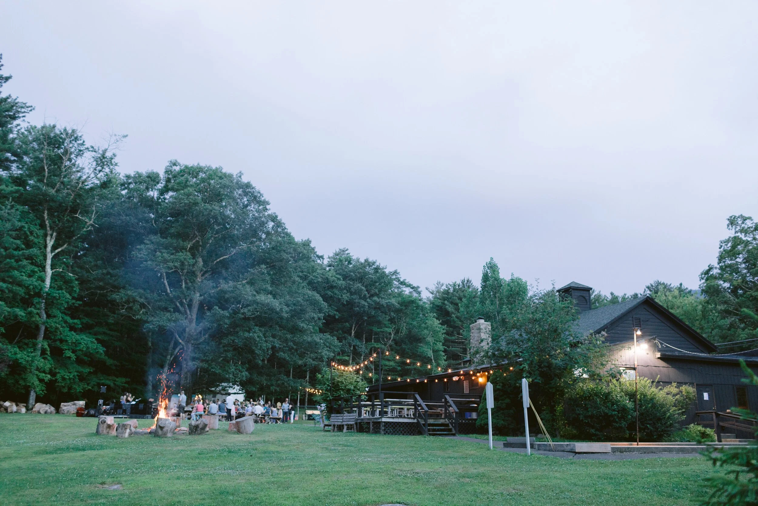 Gathering outdoors at dusk with string lights, a campfire, trees, a dark-colored house, and people seated around the fire.