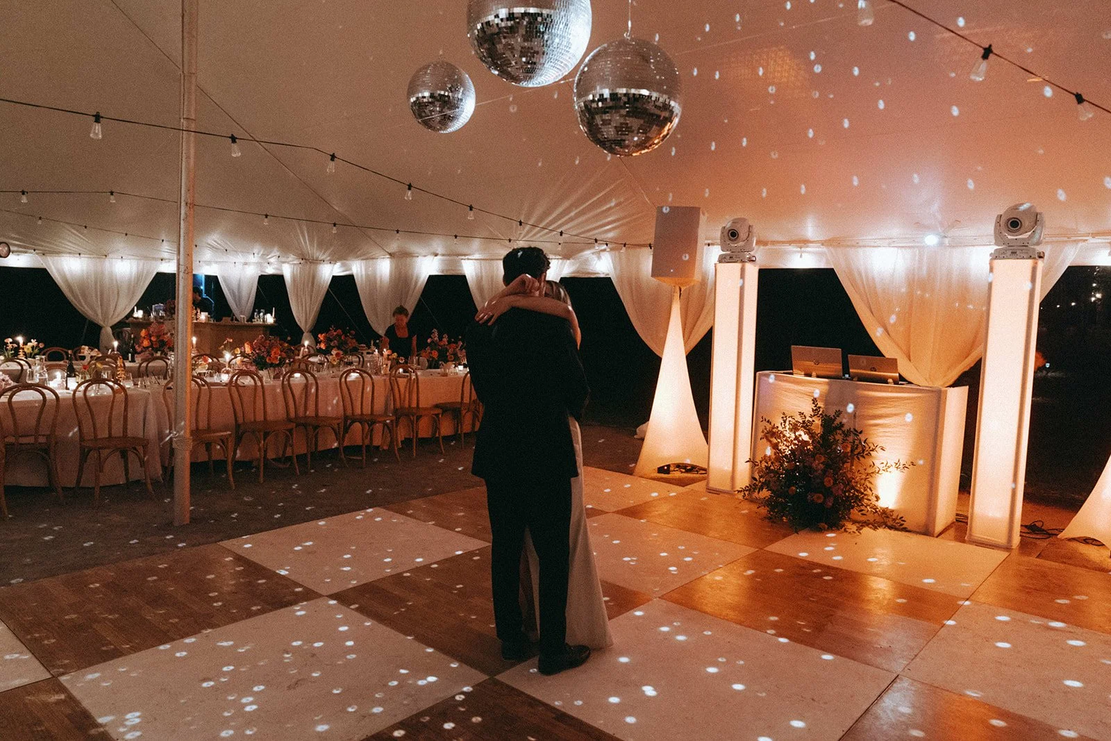 A couple dancing at a wedding reception in a decorated tent with string lights and disco balls, surrounded by tables with floral centerpieces.