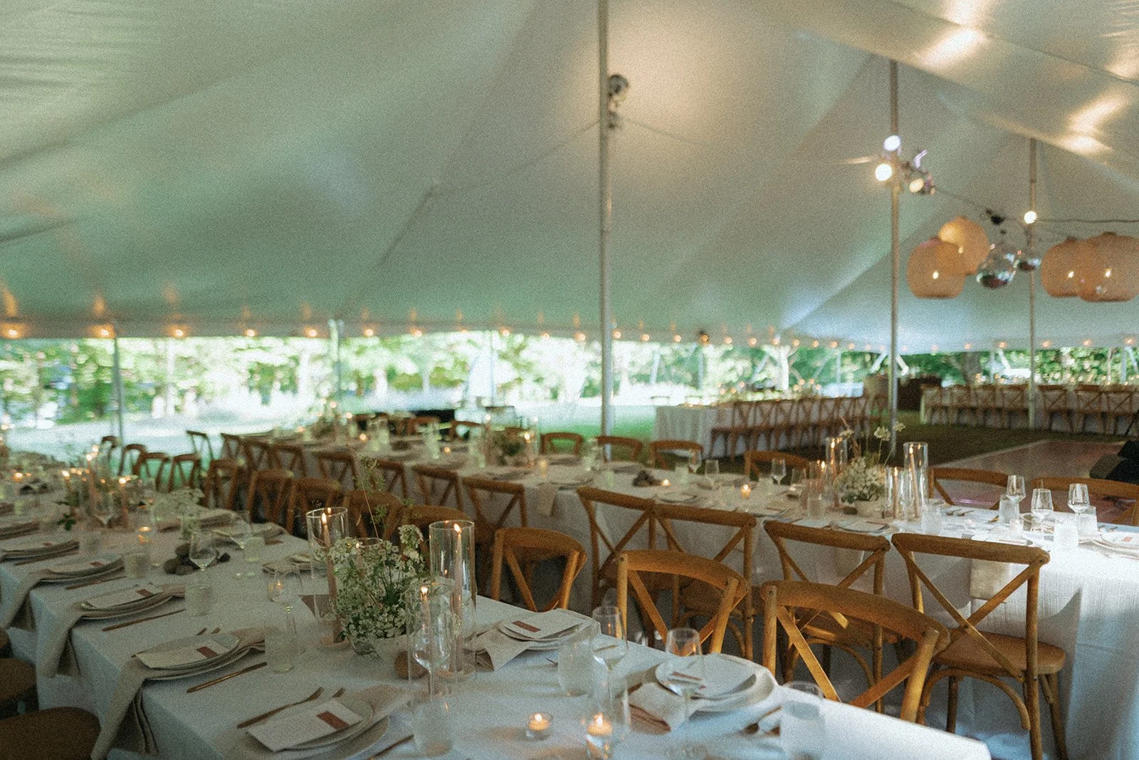 Set up for a wedding reception under a large white tent with long tables decorated with white tablecloths, candles, and floral centerpieces, surrounded by wooden chairs, with string lights and hanging paper lanterns.