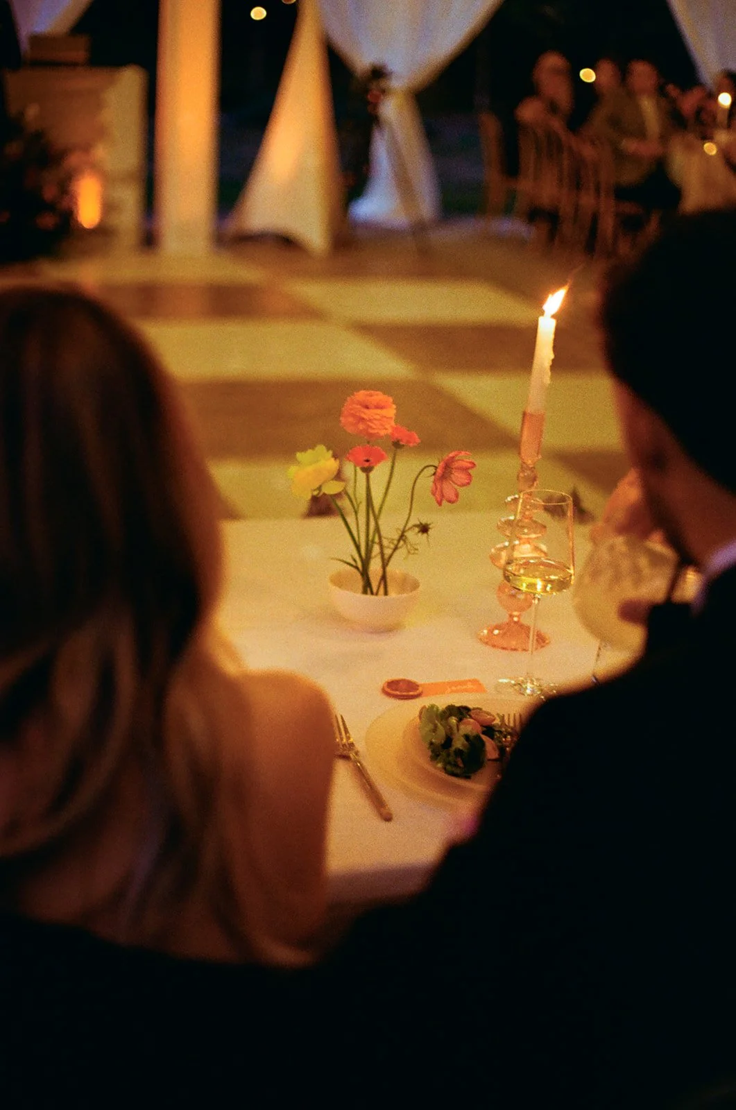A romantic dinner scene with a lit candle, a glass of white wine, a small floral arrangement, and two people sitting at a table in a softly lit outdoor setting.