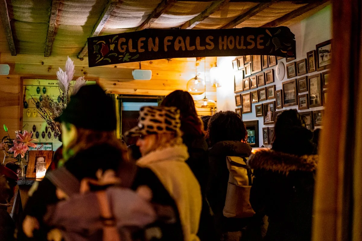 A group of people inside a cozy wooden room with a sign reading 'Glen Falls House.' The room has framed pictures on the wall, a flower arrangement on a table, and warm lighting.