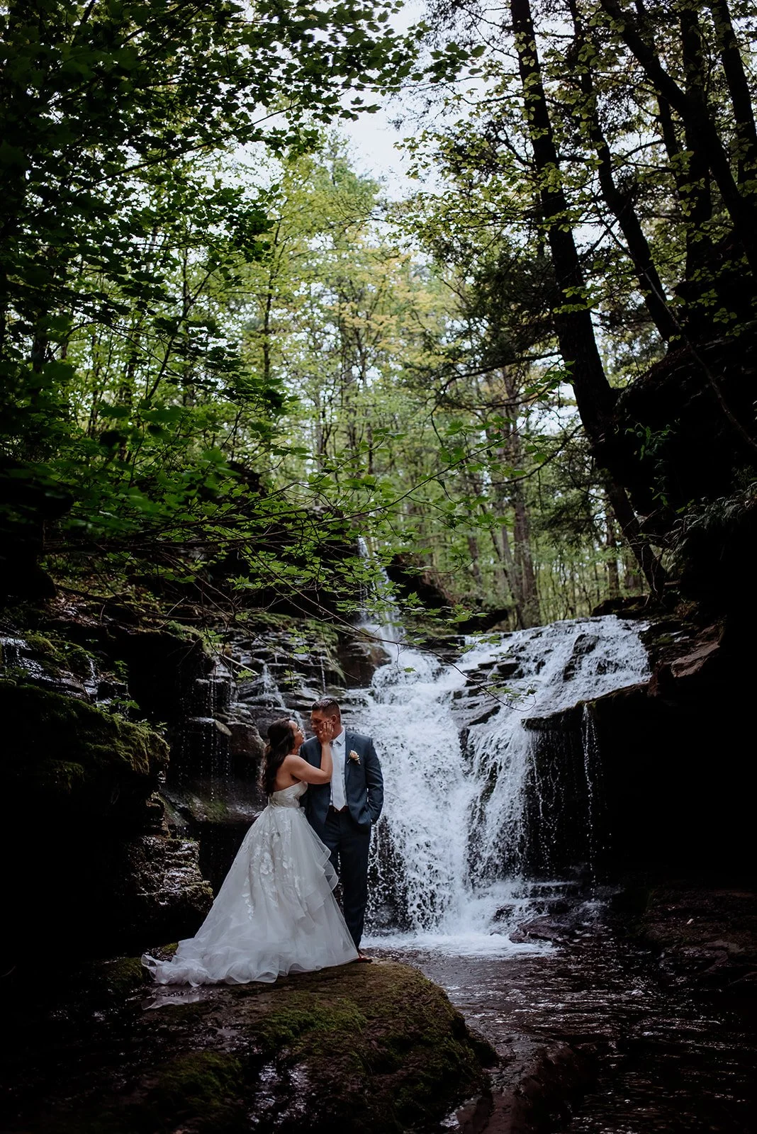 A bride and groom standing on a mossy rock in front of a small waterfall surrounded by dense green trees in a forest setting.