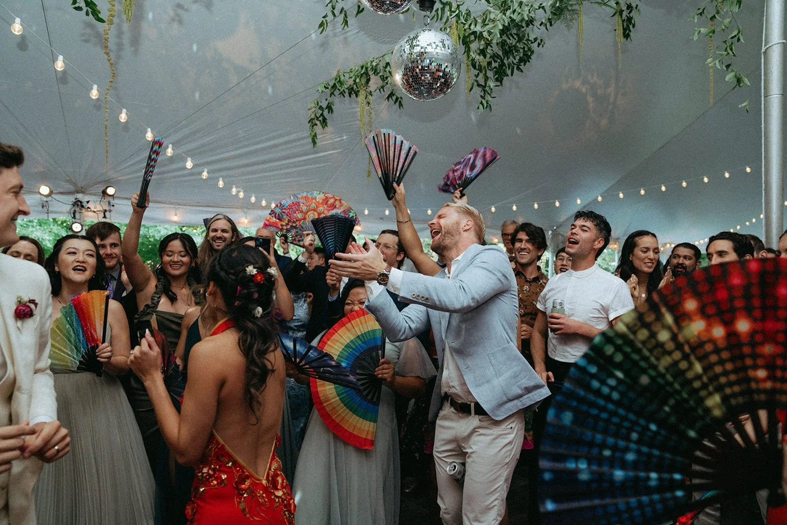 People celebrating at a party under a tent, with some holding colorful fans and a disco ball hanging from the ceiling, while enjoying and laughing.