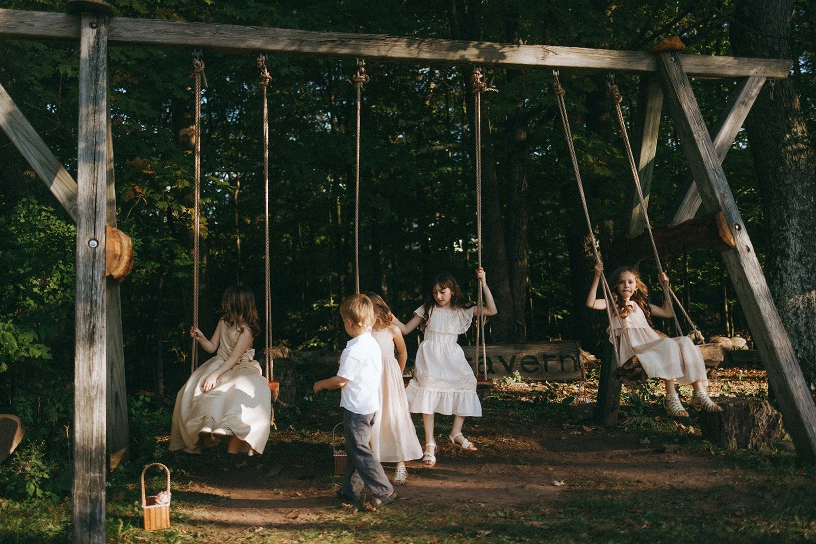Children in white dresses and a boy in a white shirt and gray pants playing on a wooden swing set in a forested area during daytime.