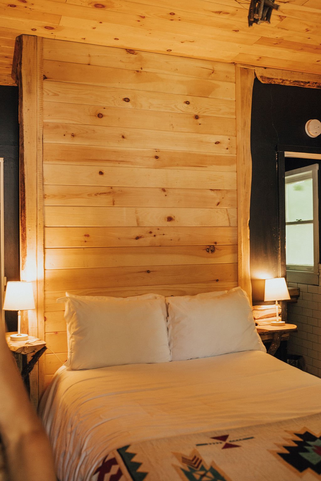A cozy bedroom with a rustic wooden headboard, cream pillows and bedspread, two bedside lamps, and a window with light coming through.