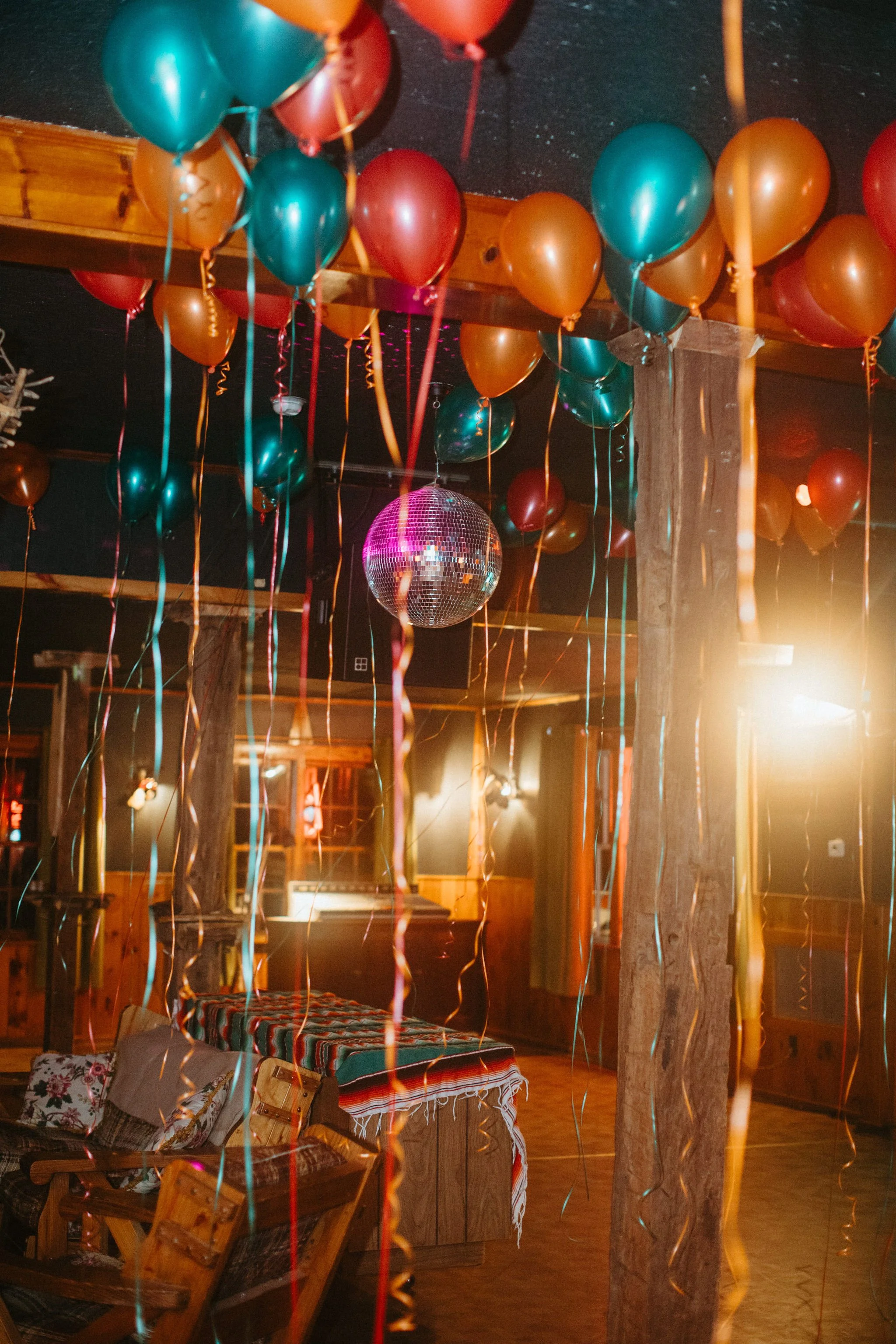 Decorated indoor space with colorful balloons and a disco ball hanging from the ceiling, warm lighting, and rustic wooden furniture.