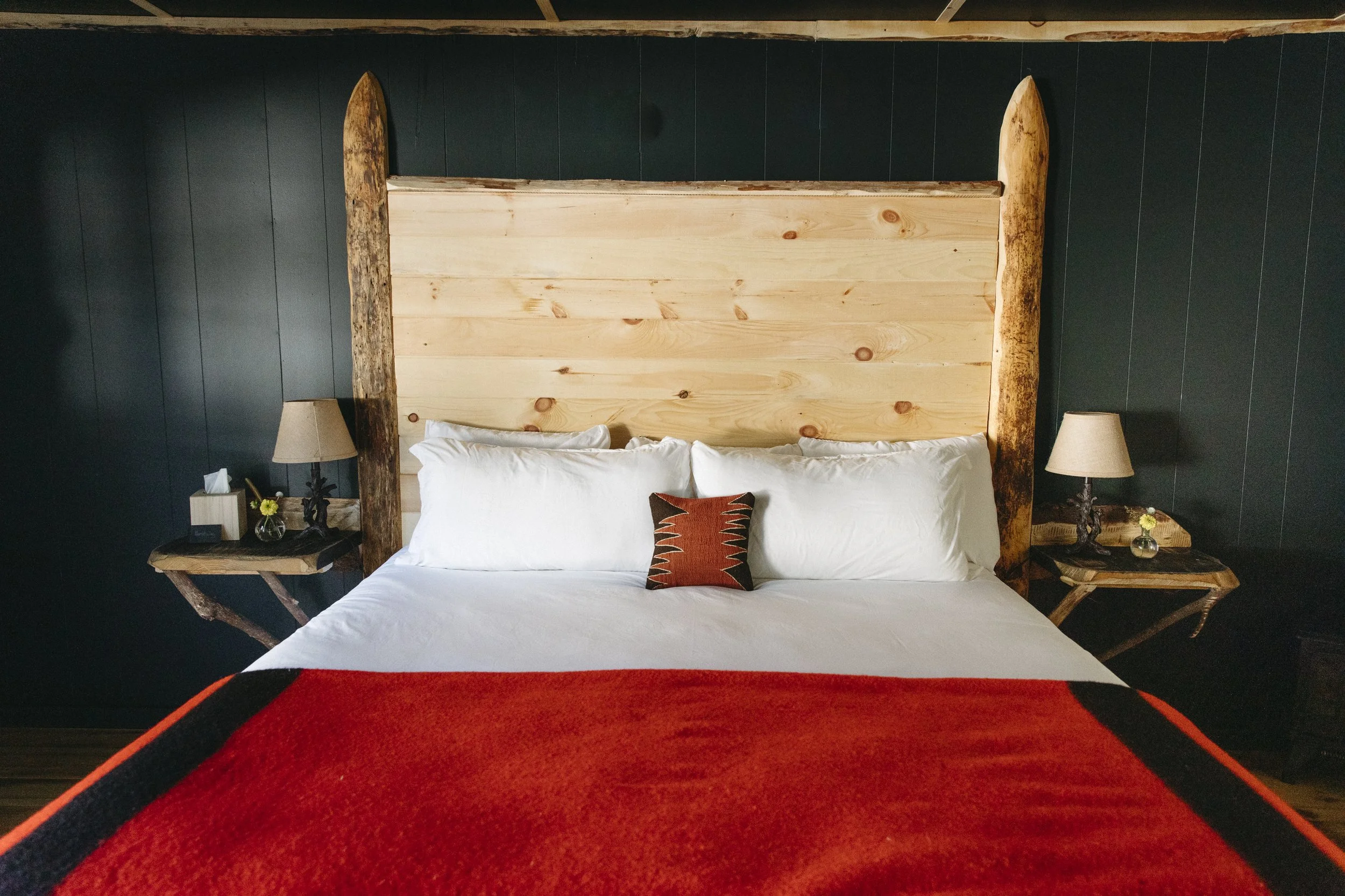 A neatly made bed with white sheets and pillows, a small decorative pillow in the middle, a wooden headboard, and dark walls. There are two nightstands with lamps and small decorative items on each side of the bed.