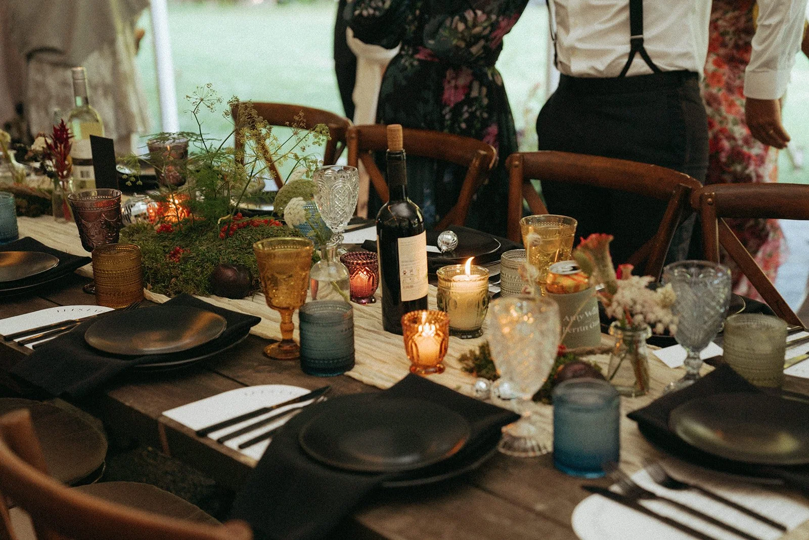 A rustic dining table decorated for a special occasion with black plates, amber and blue glasses, candles, floral arrangements, and a bottle of wine. People dressed semi-formally stand around the table.