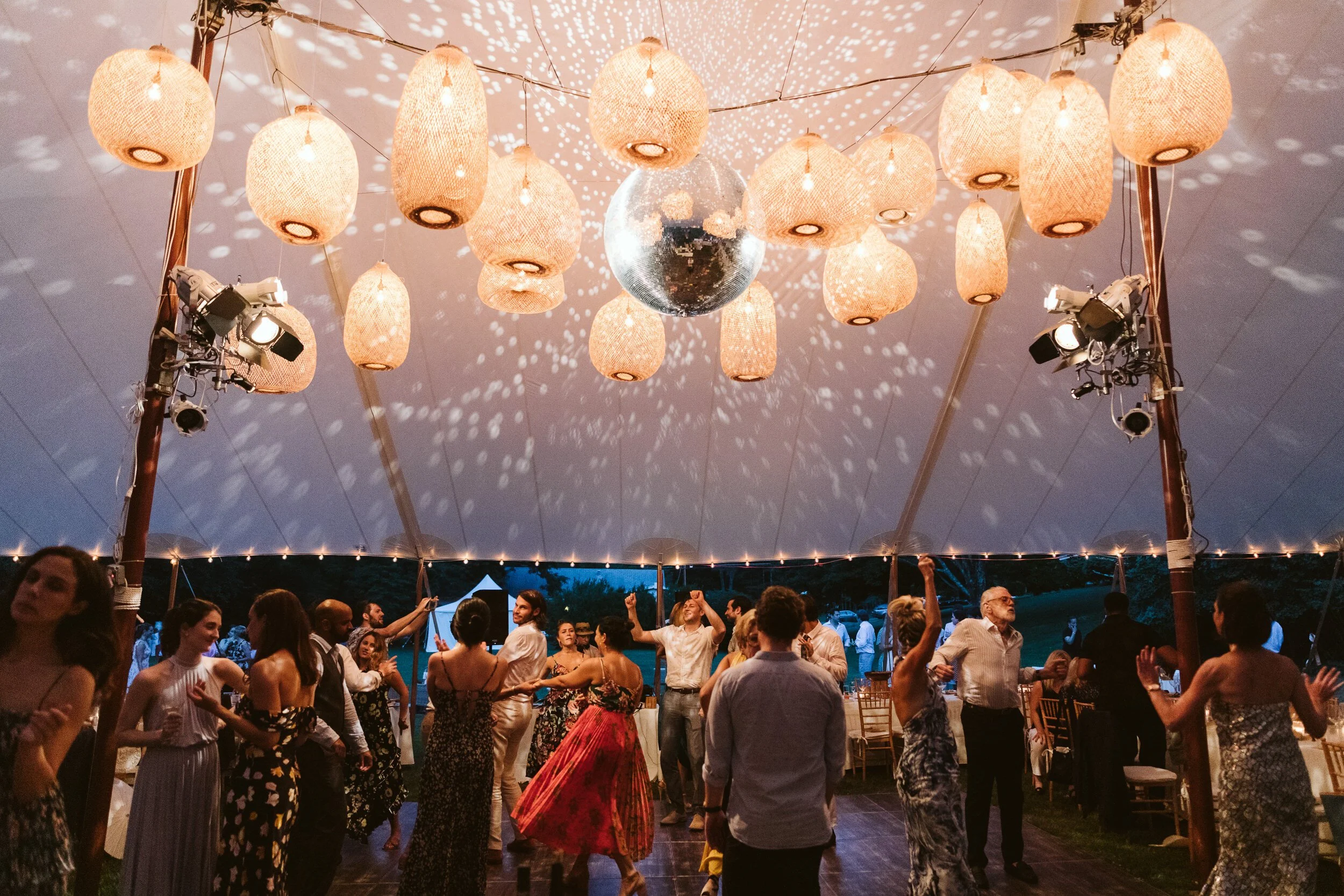 People dancing under a large tent with hanging paper lanterns and a disco ball, surrounded by string lights.
