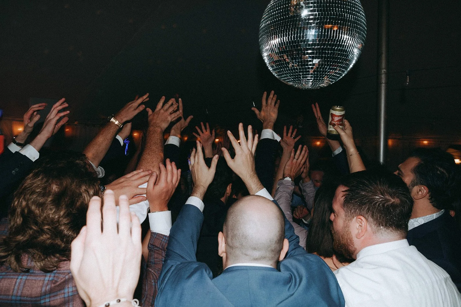 Crowd of people dancing and raising hands in a party with a disco ball overhead, some holding drinks.