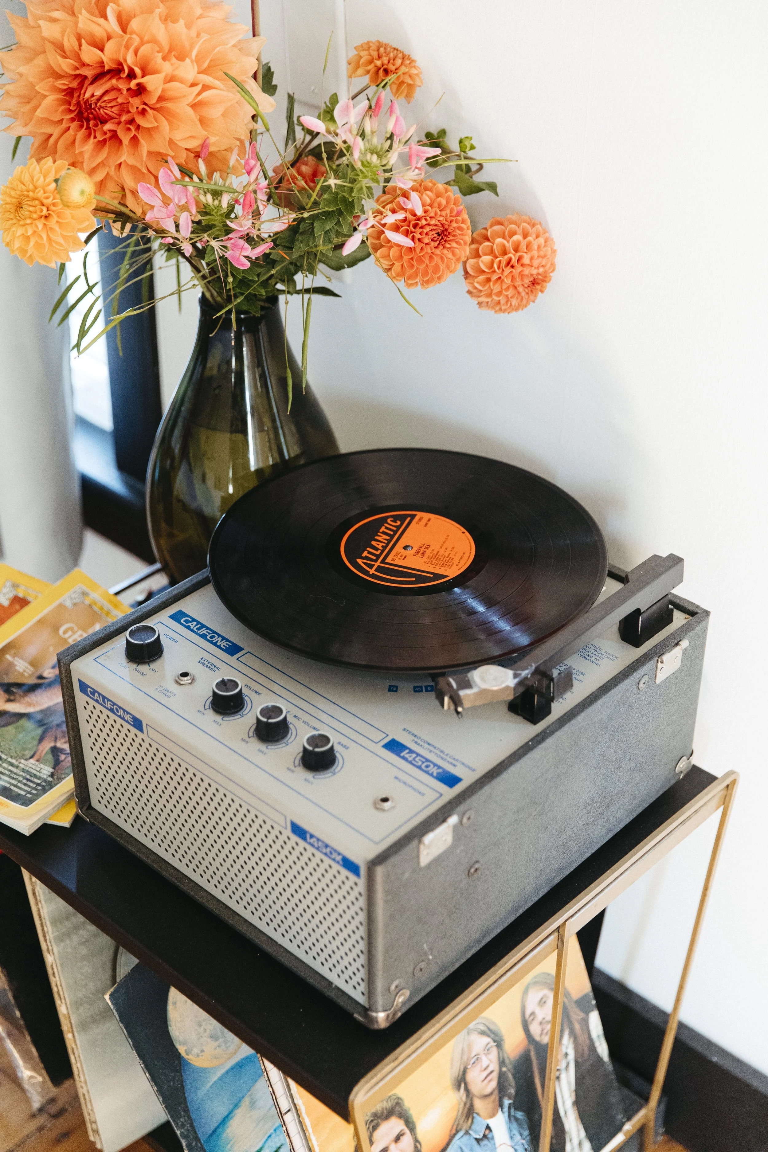 A vintage record player on a black side table with a black vinyl record on the turntable, positioned near a large glass vase filled with orange and pink flowers.