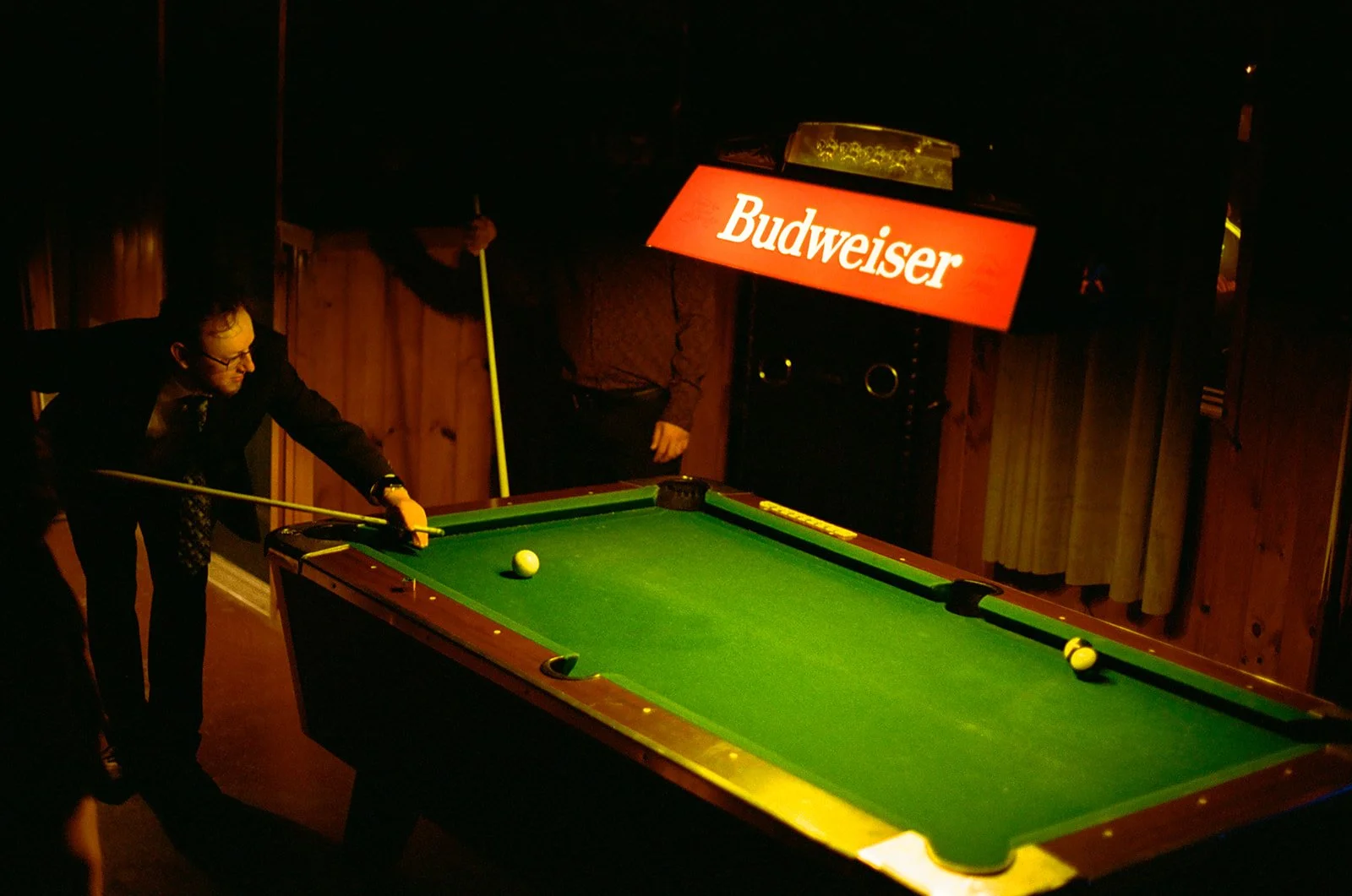 A man in a suit aiming a cue at a pool ball on a green billiards table in a dimly lit bar or game room, with a Budweiser sign hanging above the table.