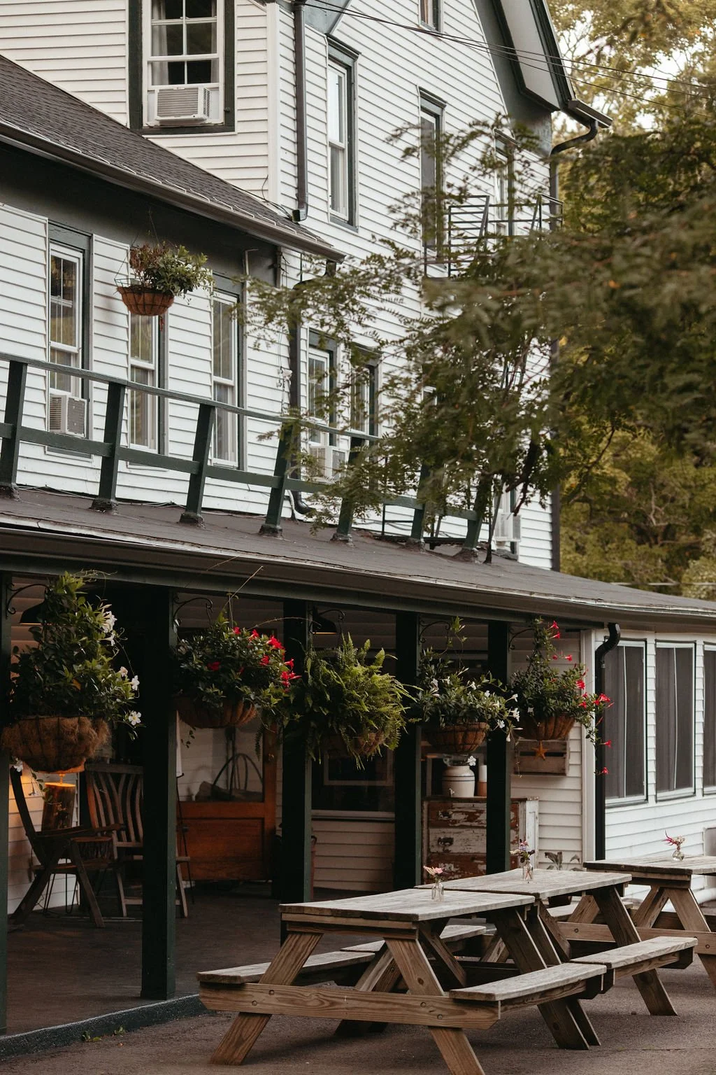 The image shows a patio area of a white house with black trim, featuring hanging flower baskets, wooden picnic tables, and a small porch with chairs and potted plants. The house has multiple windows, some with air conditioning units, and a small balc