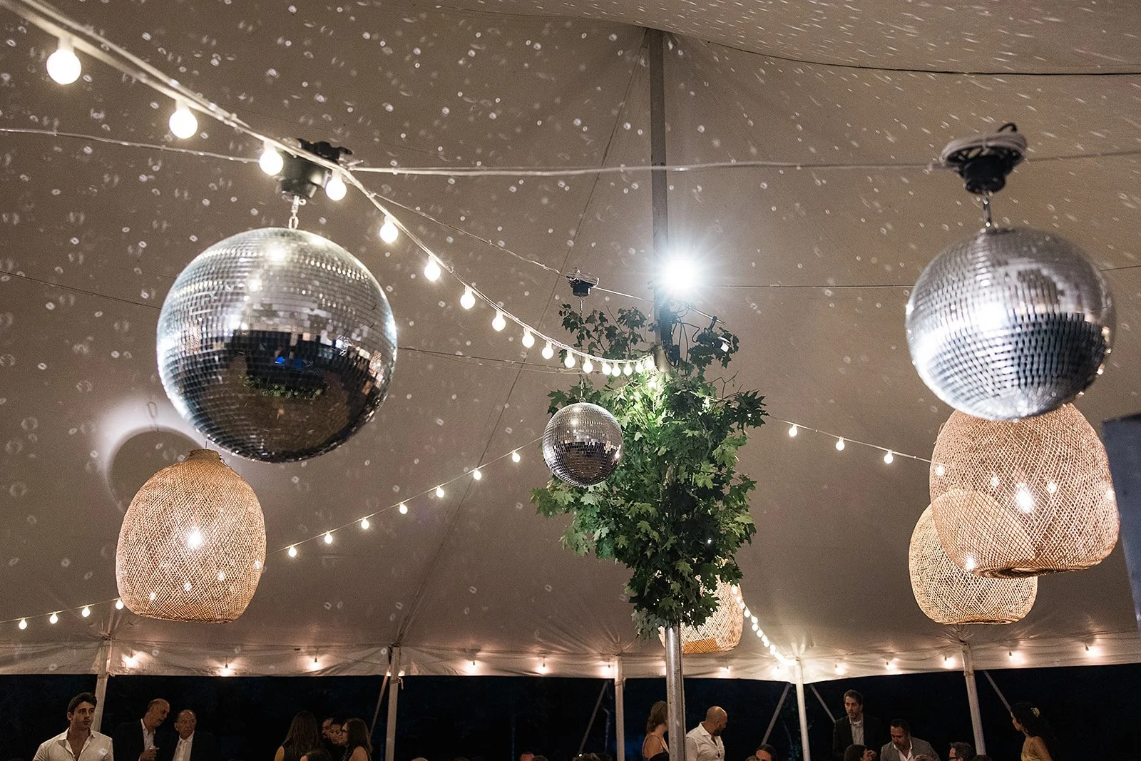 Decorative disco balls and woven lanterns hanging from string lights under a tent ceiling at a nighttime event with people in the background.