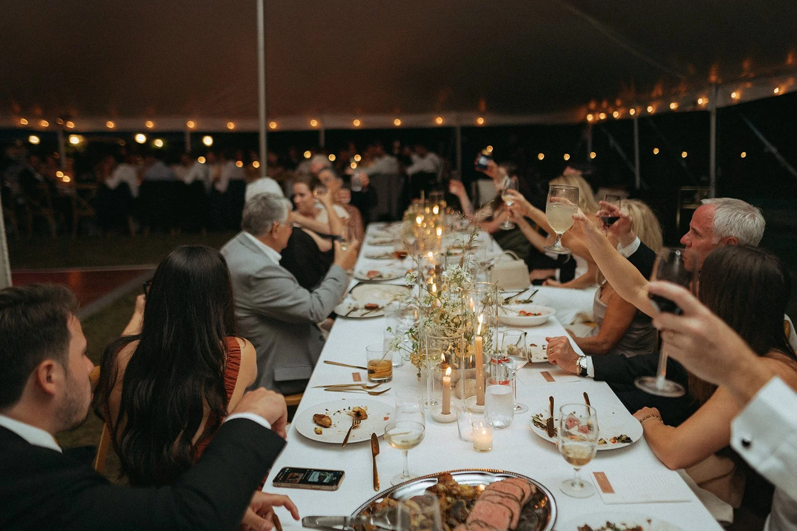People gather around a long decorated table at an outdoor evening event, raising glasses for a toast under a tent with string lights.