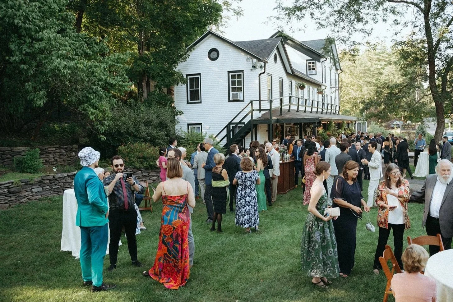 Outdoor gathering of people in front of a white house, socializing on a lawn with some tables and decorations, under trees with a two-story house in the background.