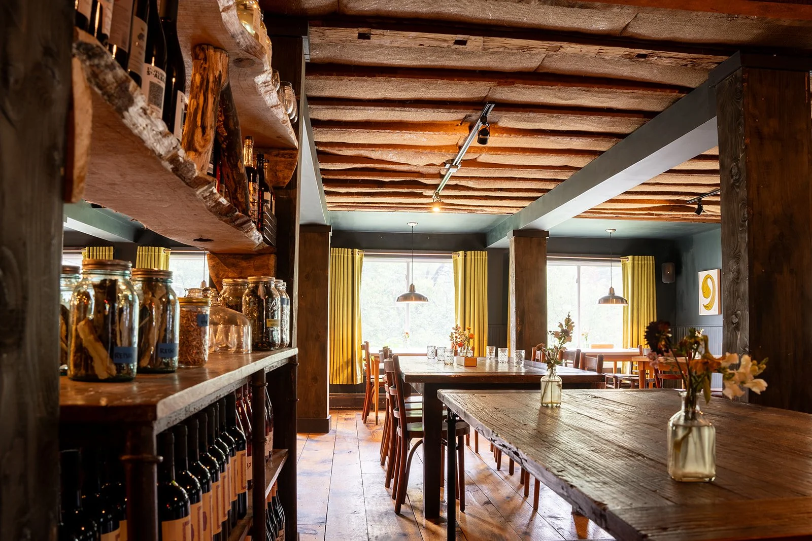Inside a cozy restaurant with wooden beams on the ceiling, large windows with yellow curtains, and wooden tables decorated with small vases of flowers.